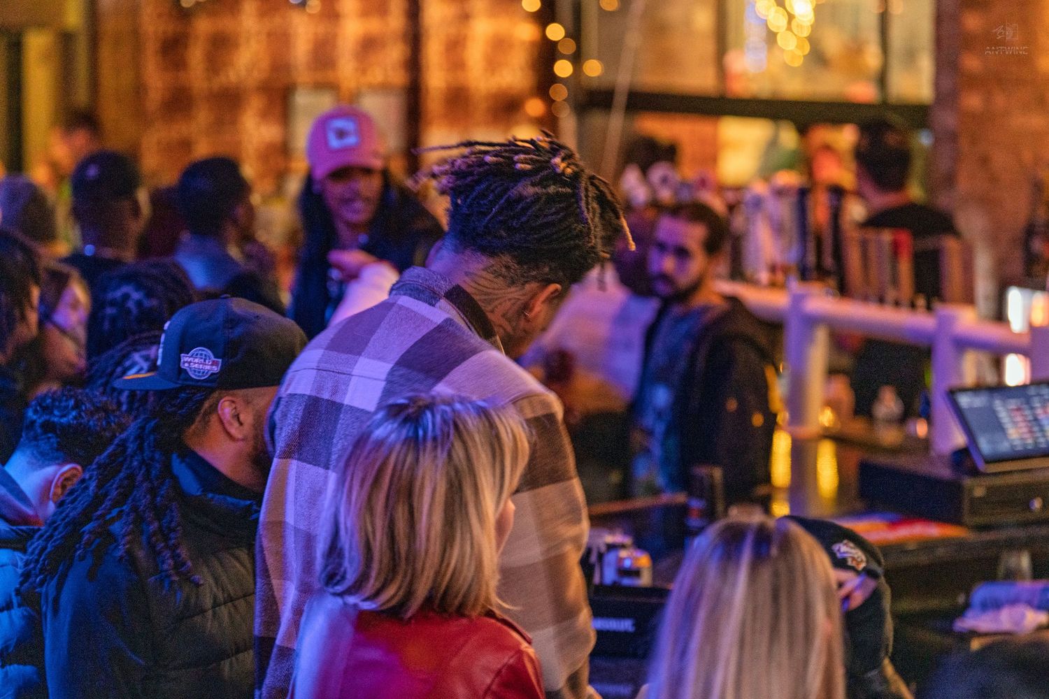 Man with dreadlocks in plaid shirt mingles at an outdoor bar event with warm string lights.