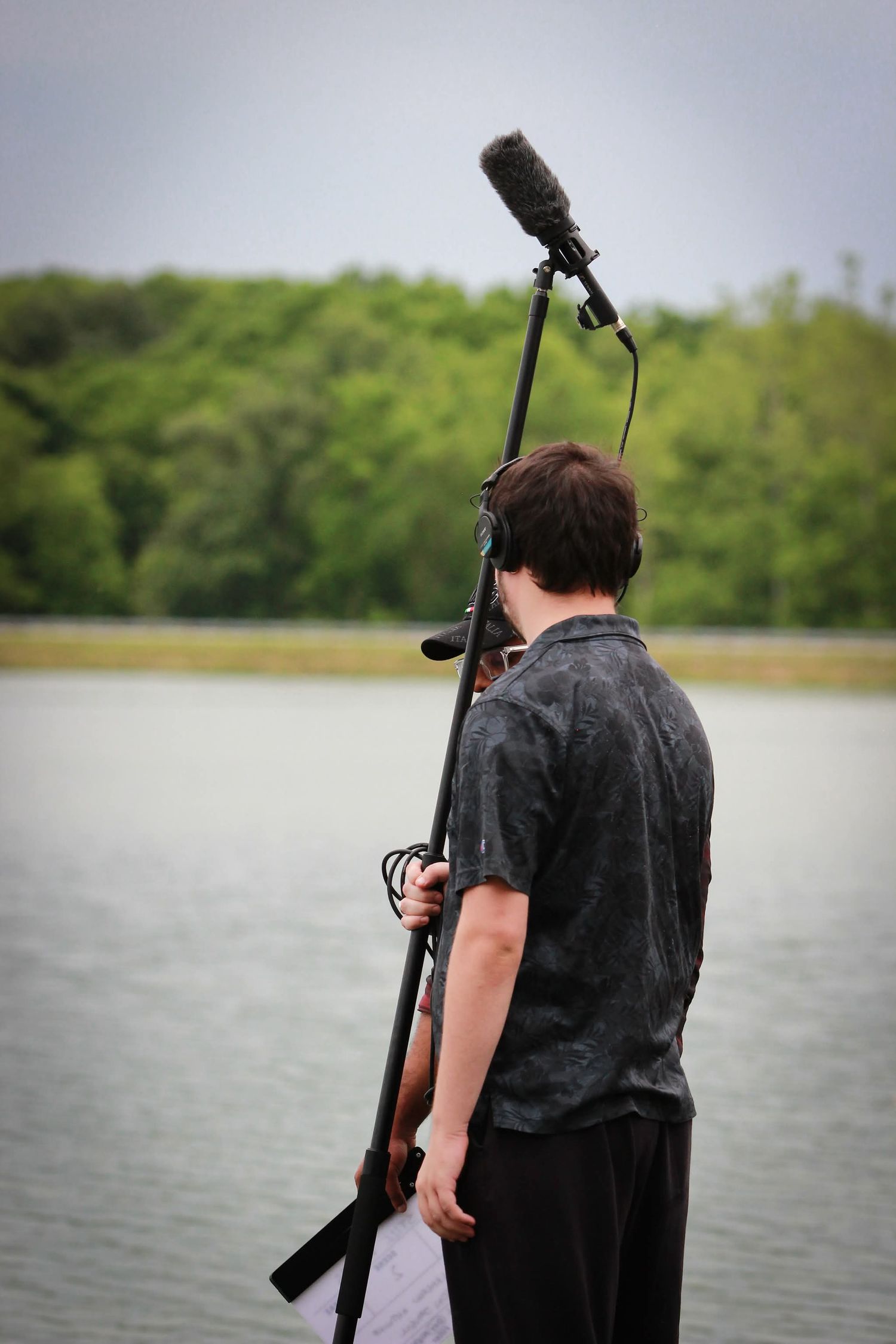 A boom mic operator holds a shotgun microphone on a pole near a serene lake with lush green trees in the background.