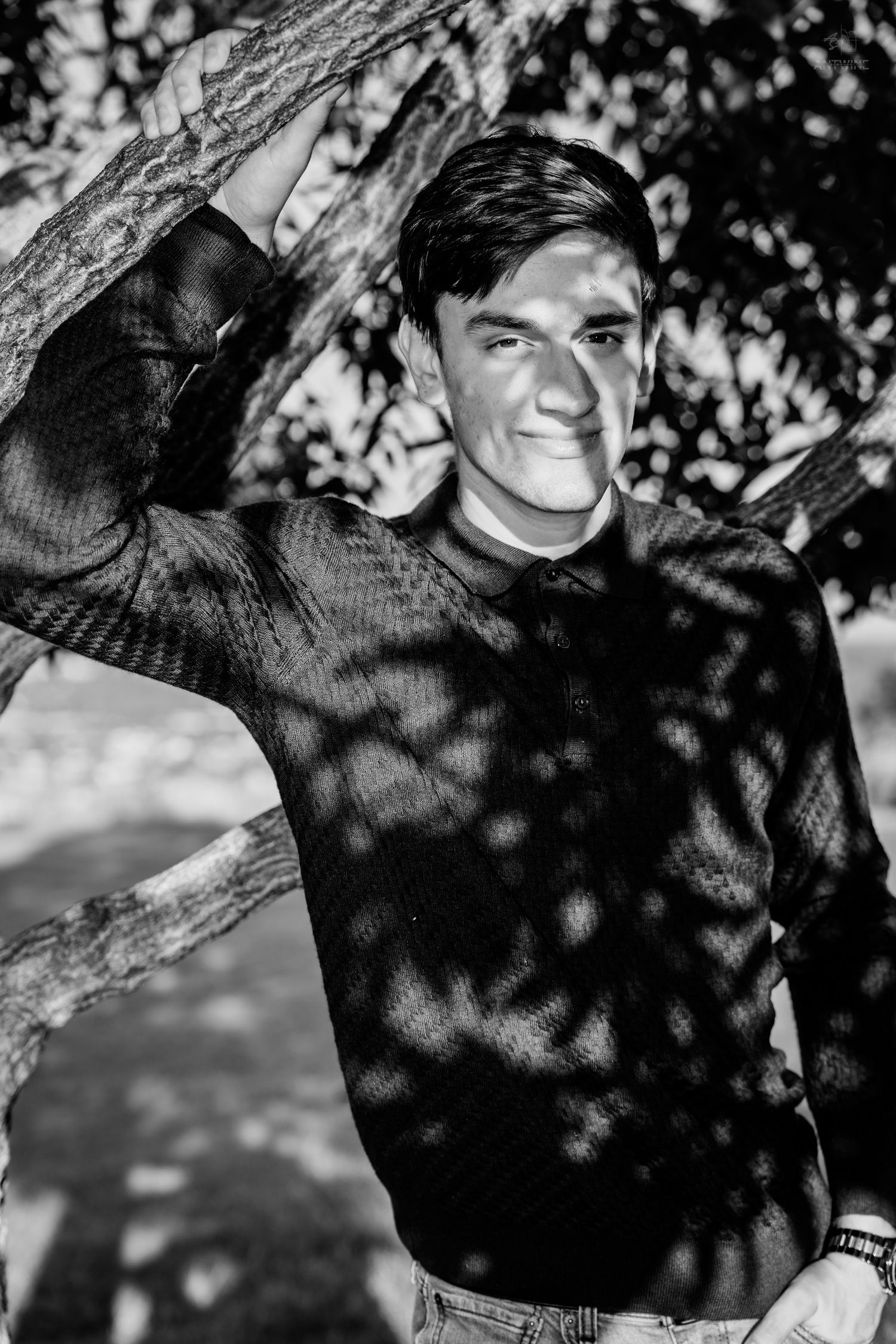 Young man smiling beside a tree, wearing a dark sweater, with dappled shadows in a black and white outdoor portrait.