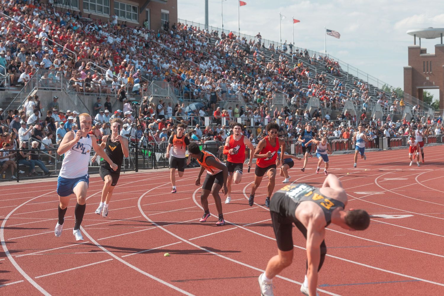 Large group of runners compete in track race before packed stadium crowd at outdoor track and field meet.