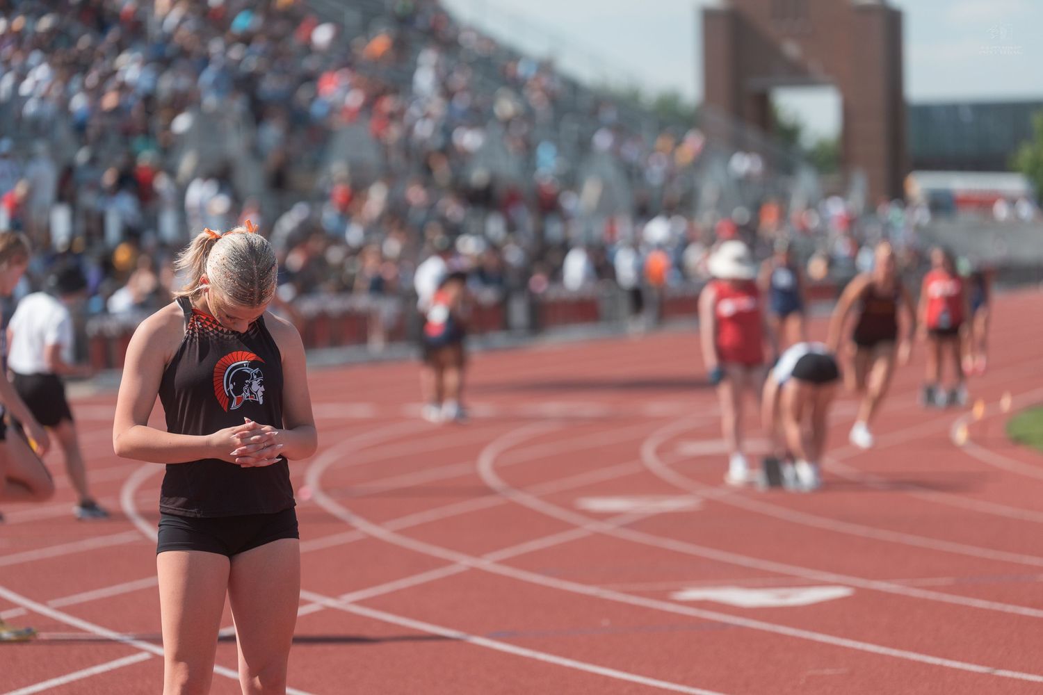 Female track athlete in black uniform checks her phone on a red track during a busy outdoor meet.