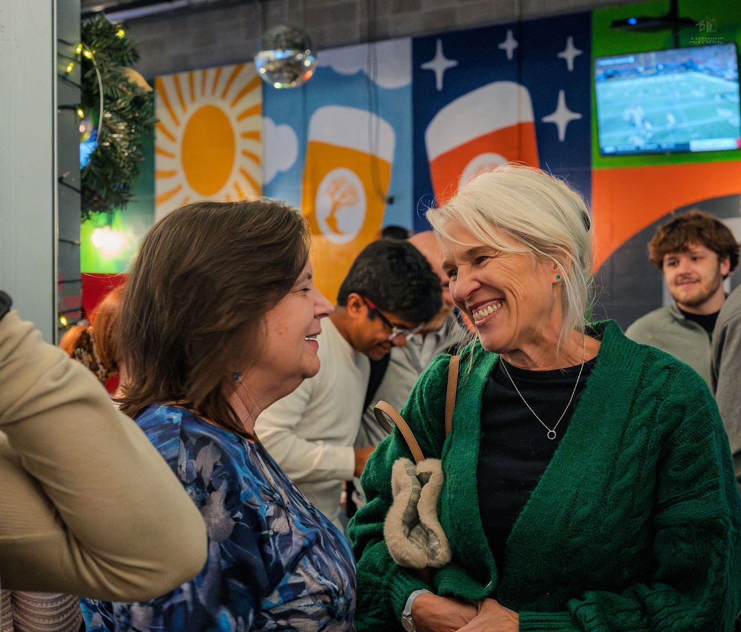 Two women laughing and talking together at a lively indoor social event.