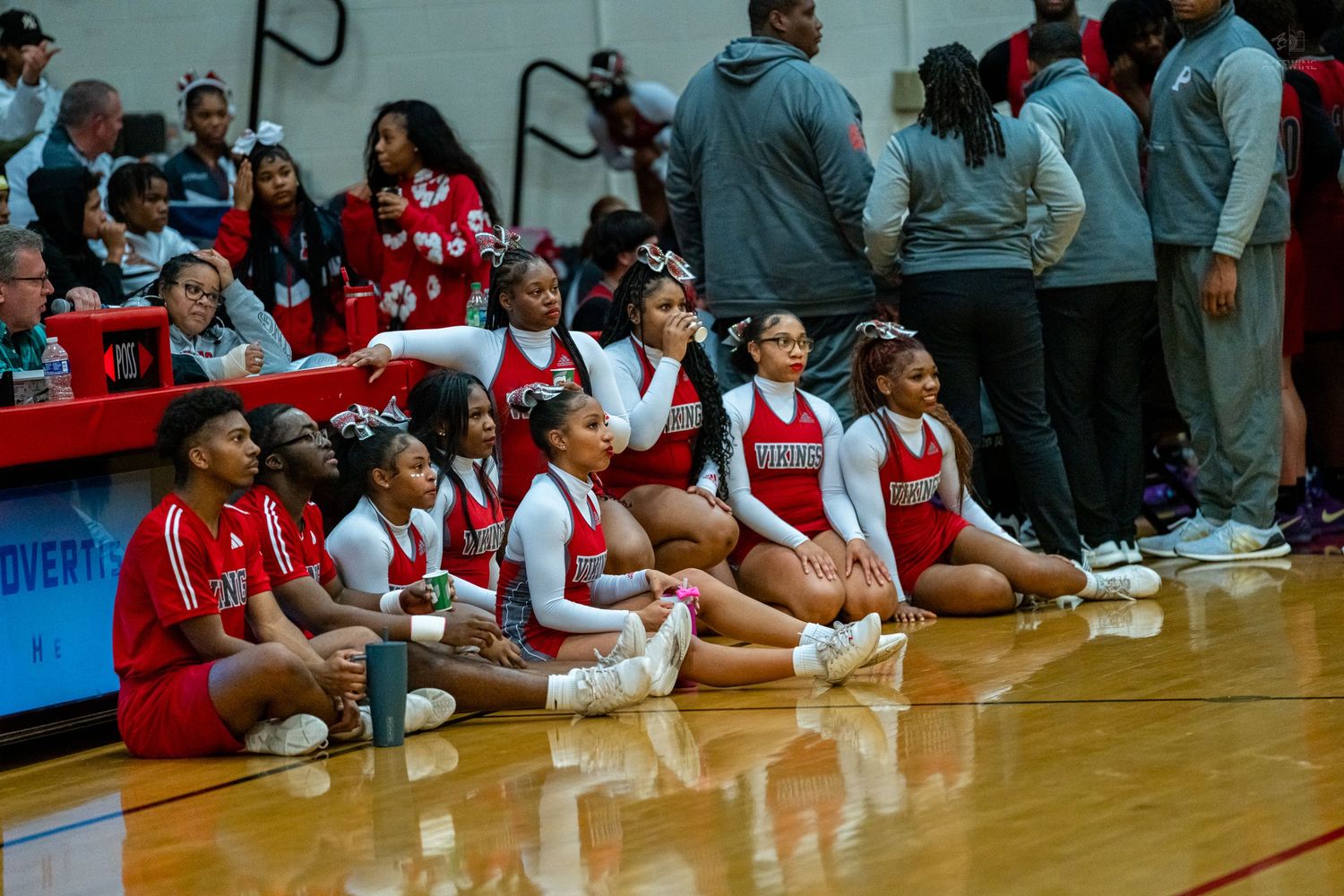 High school cheerleaders sit on the hardwood court floor during a basketball game sideline break.