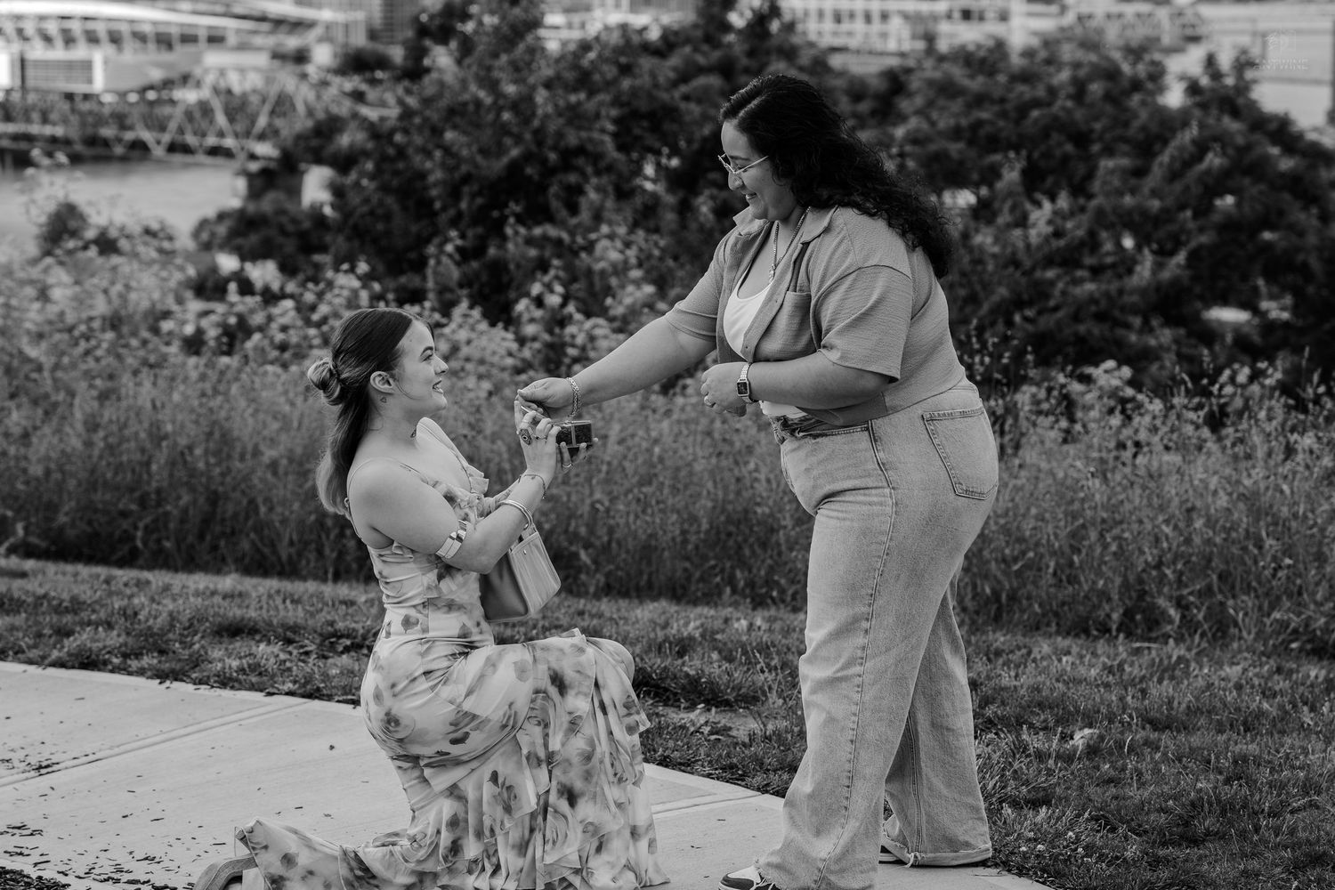 Woman kneeling on sidewalk proposing to partner in black and white outdoor photo.