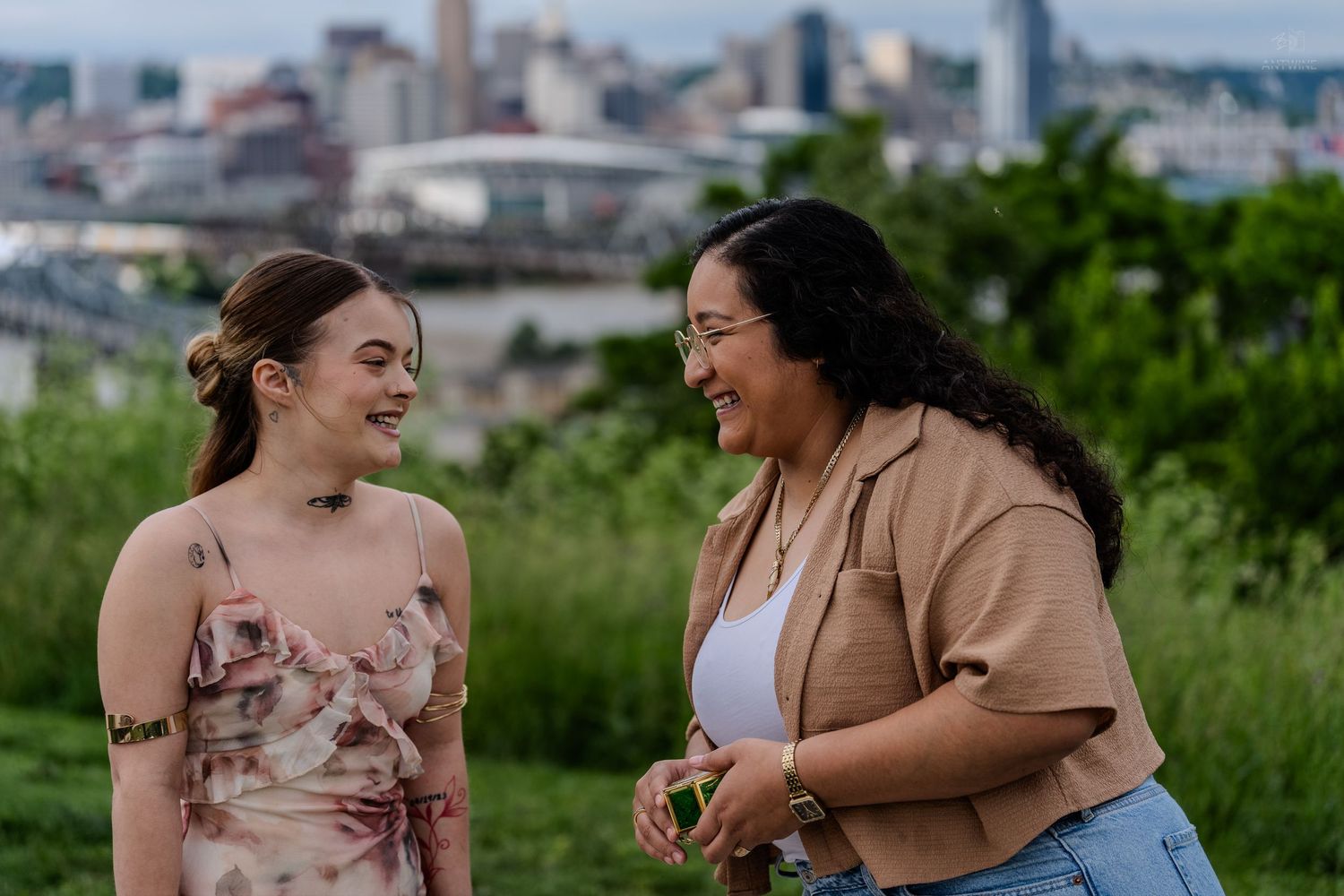 Two women laughing and talking outdoors with a city skyline in the background.