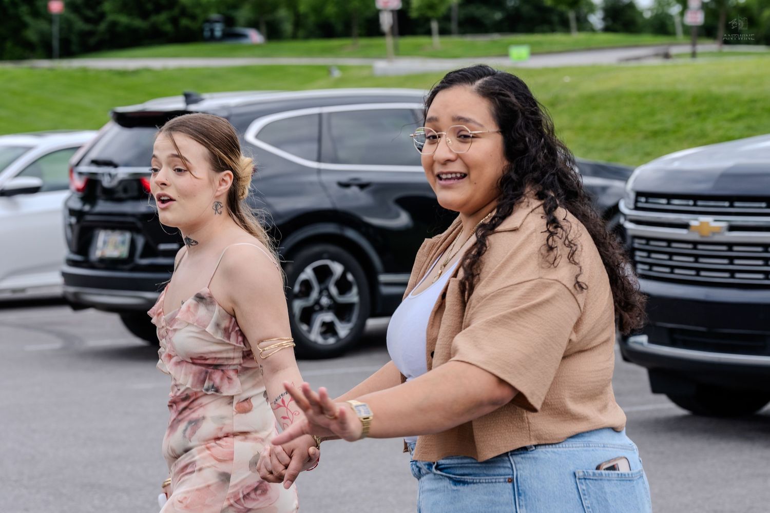 Two women laugh and hold hands while walking through a sunny parking lot together.