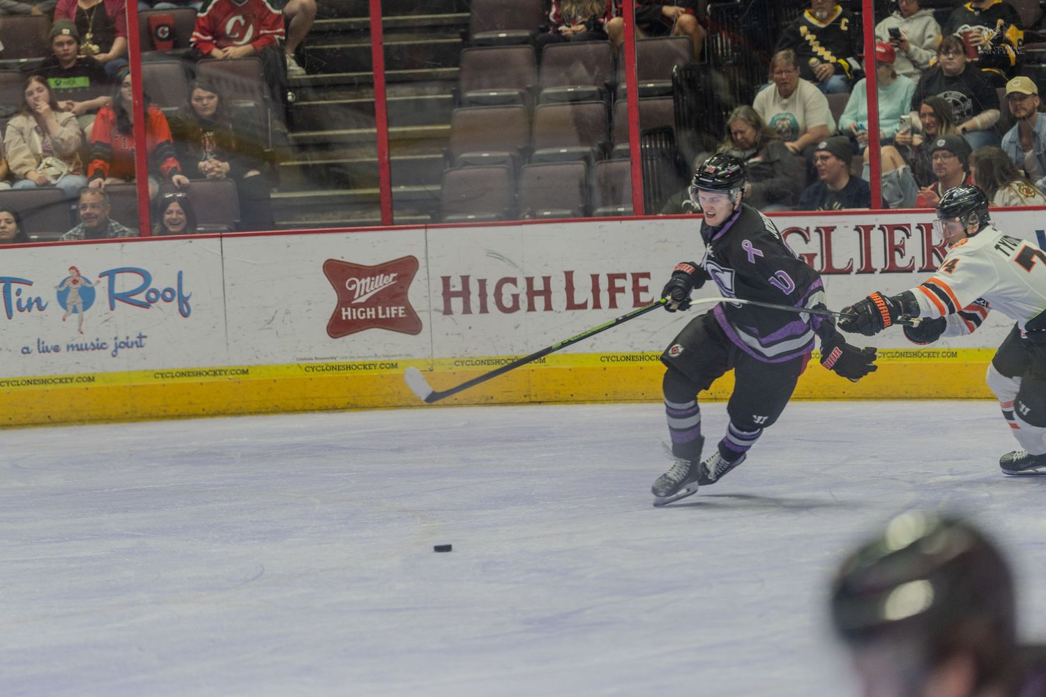 Hockey player in dark uniform shoots puck on ice with High Life and Gliers advertisements visible in background.