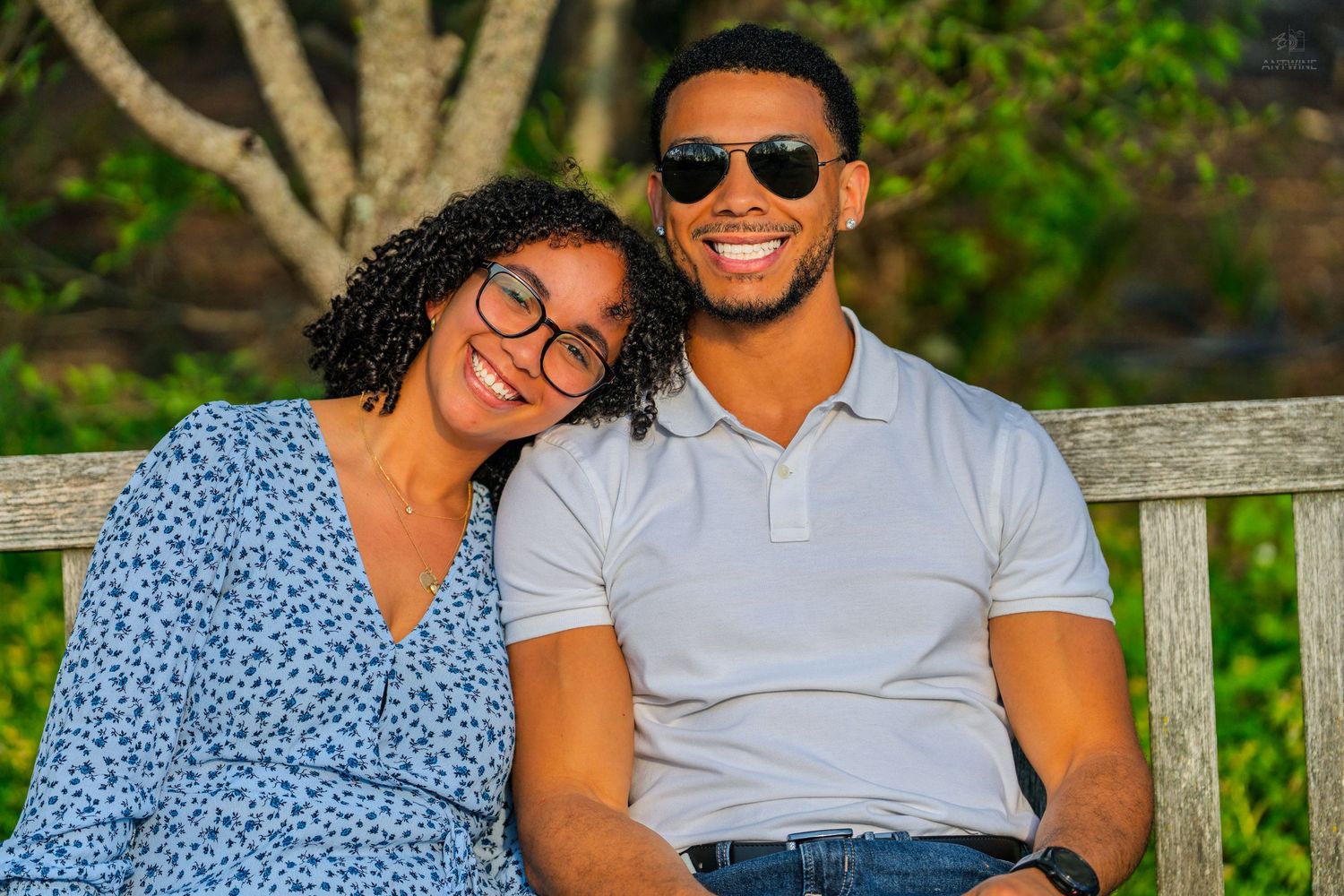 Smiling couple sits on a wooden bench outdoors in golden light, woman leaning on mans shoulder.