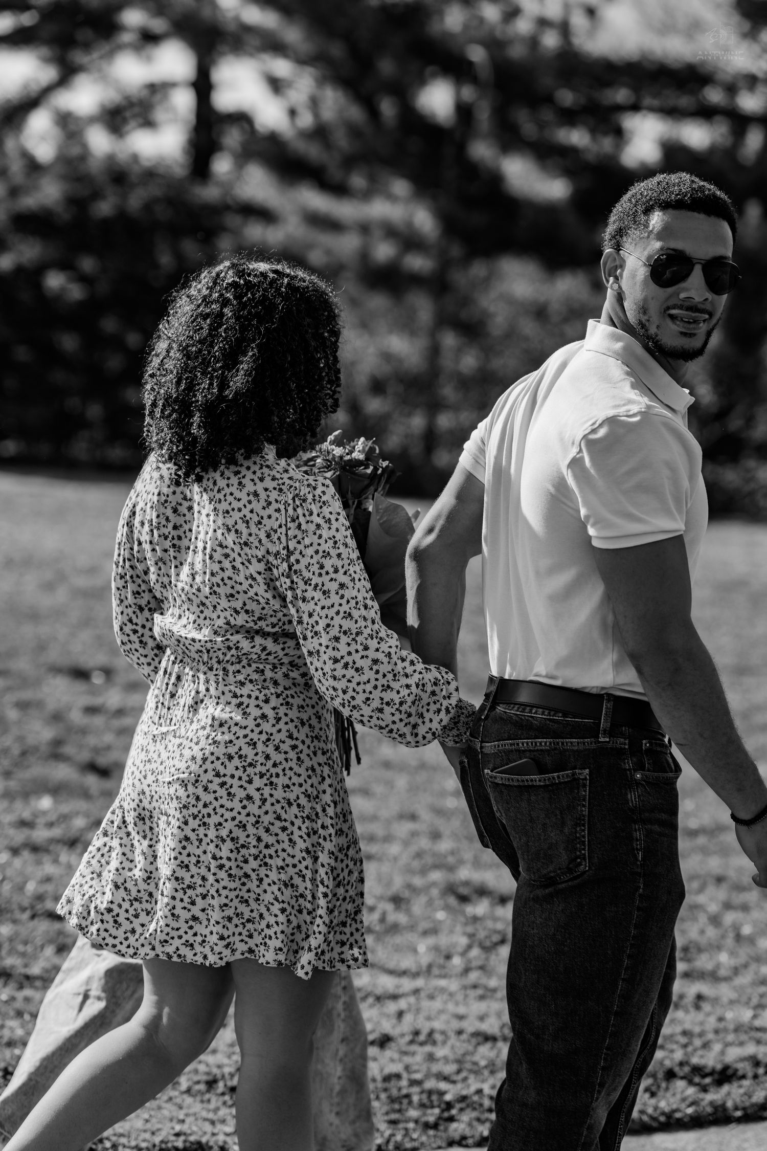 A smiling man in sunglasses glances back while walking with a woman in a floral dress in black and white.