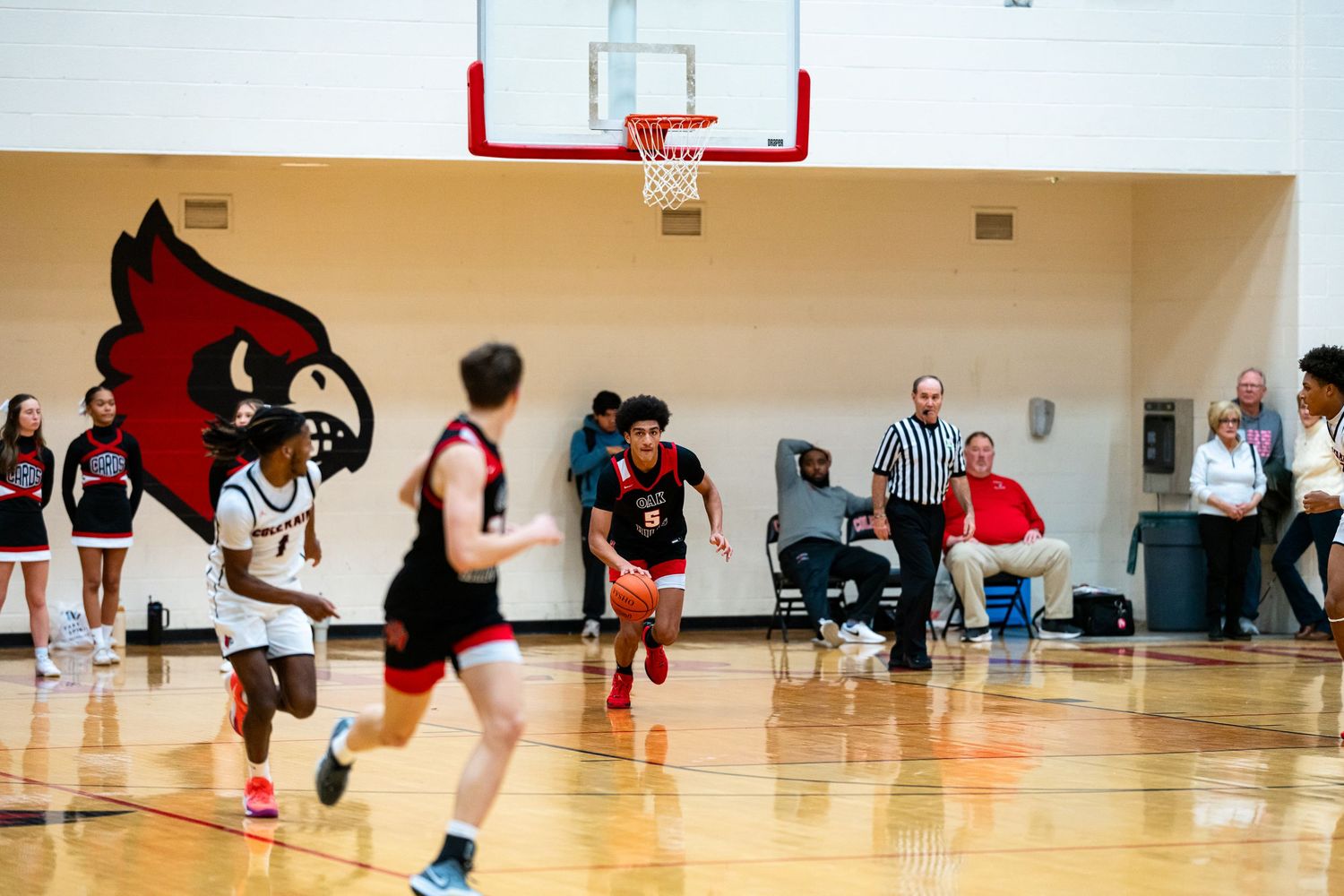 High school basketball players compete on a court featuring a red cardinal mascot logo on the wall.