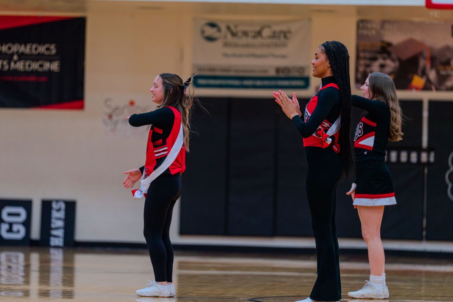Three high school cheerleaders in red and black uniforms perform a routine on a basketball court sideline.