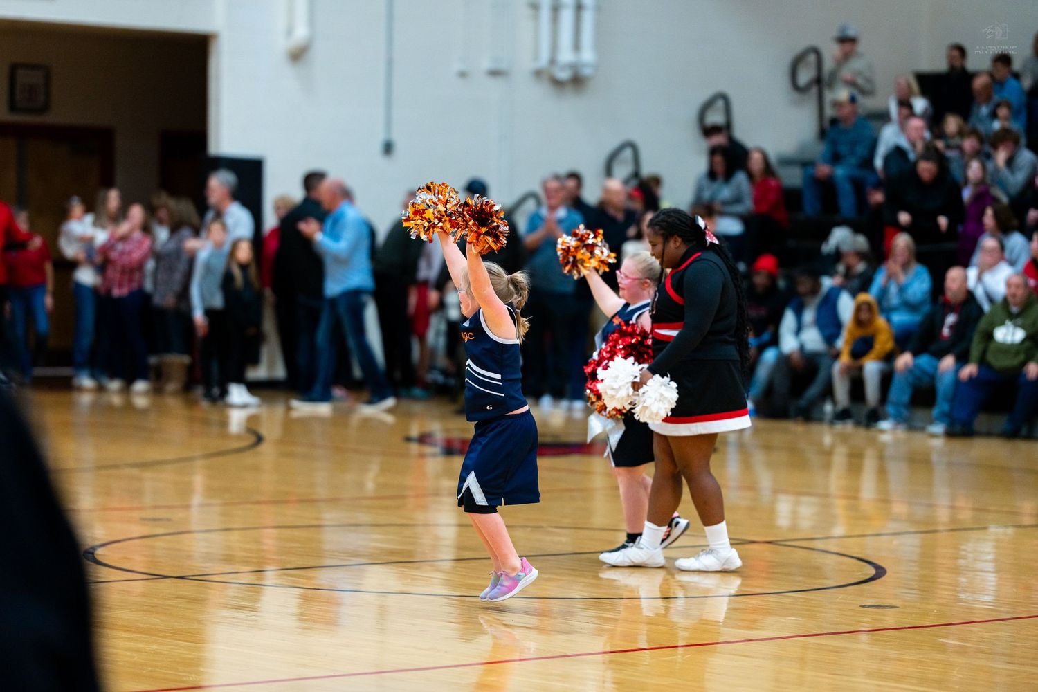 Youth and college cheerleaders perform together with colorful pom-poms on a packed basketball court.
