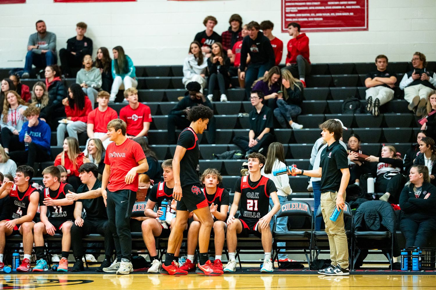 High school basketball players sit on the bench during a game in a packed gymnasium with fans in the stands.
