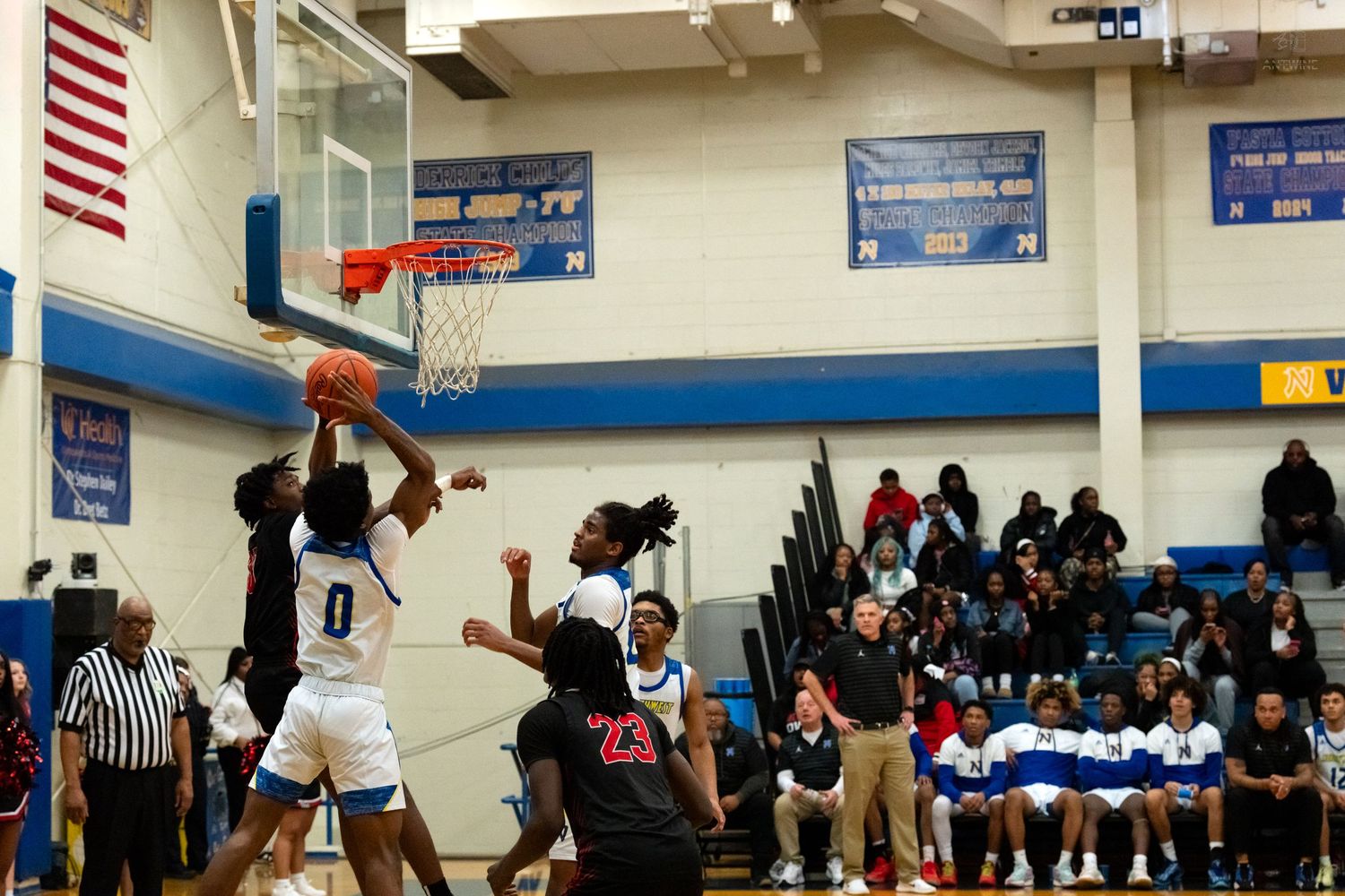 Player #0 in white drives to the basket against defenders in a packed high school basketball gym.