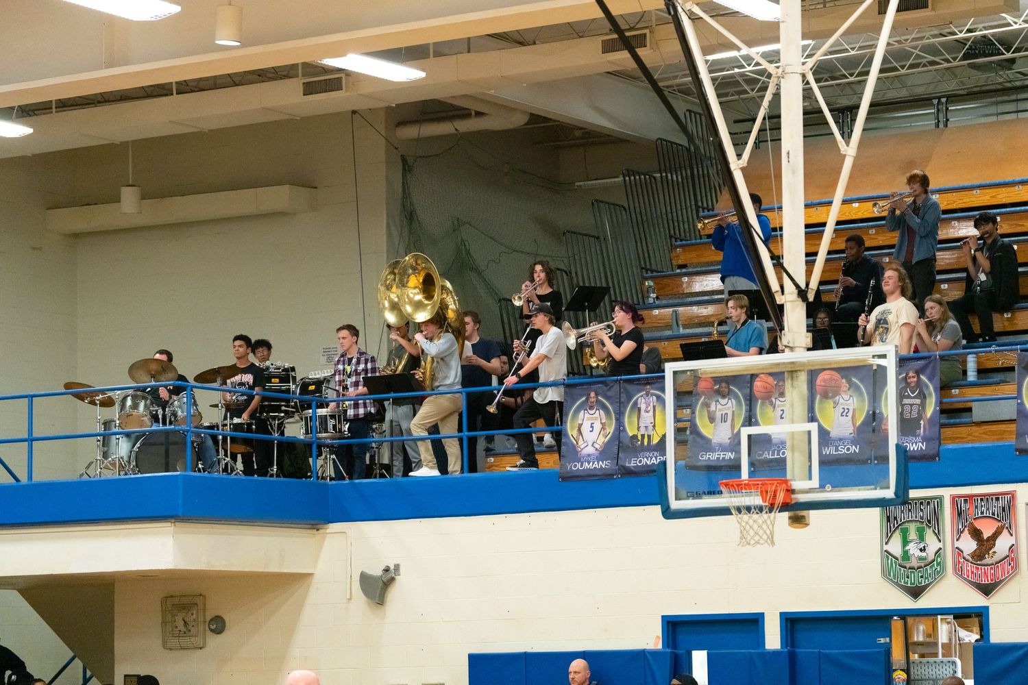 High school pep band performs from the upper balcony of a blue gymnasium during a basketball game.