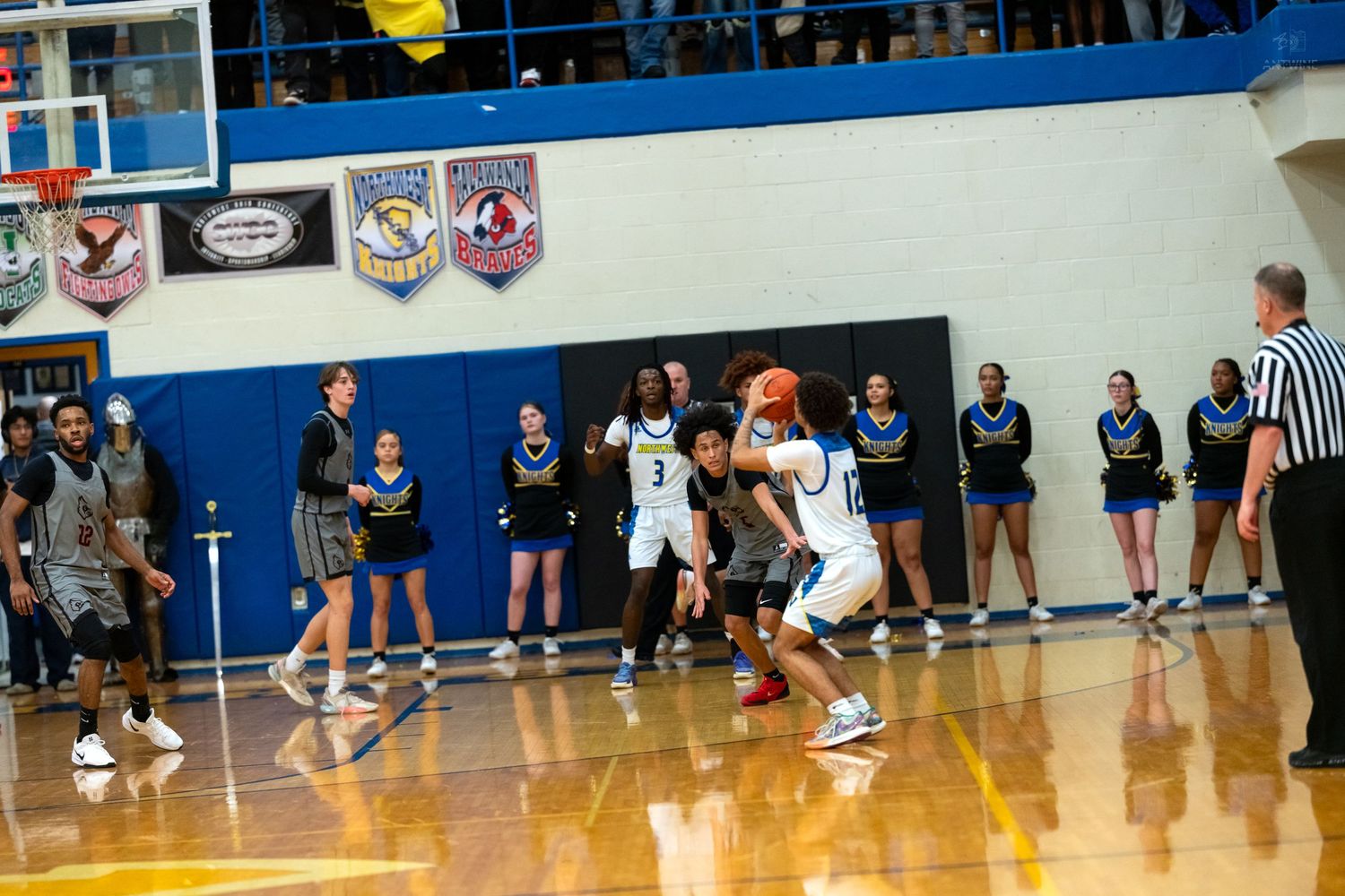 High school basketball player dribbles past defender on hardwood court with spectators and banners in background.