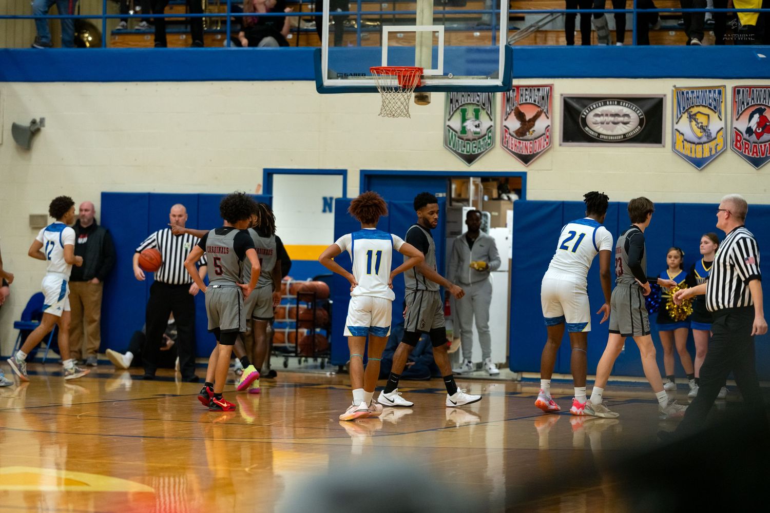 High school basketball game with players and referees on a blue and yellow gymnasium court with spectators watching.