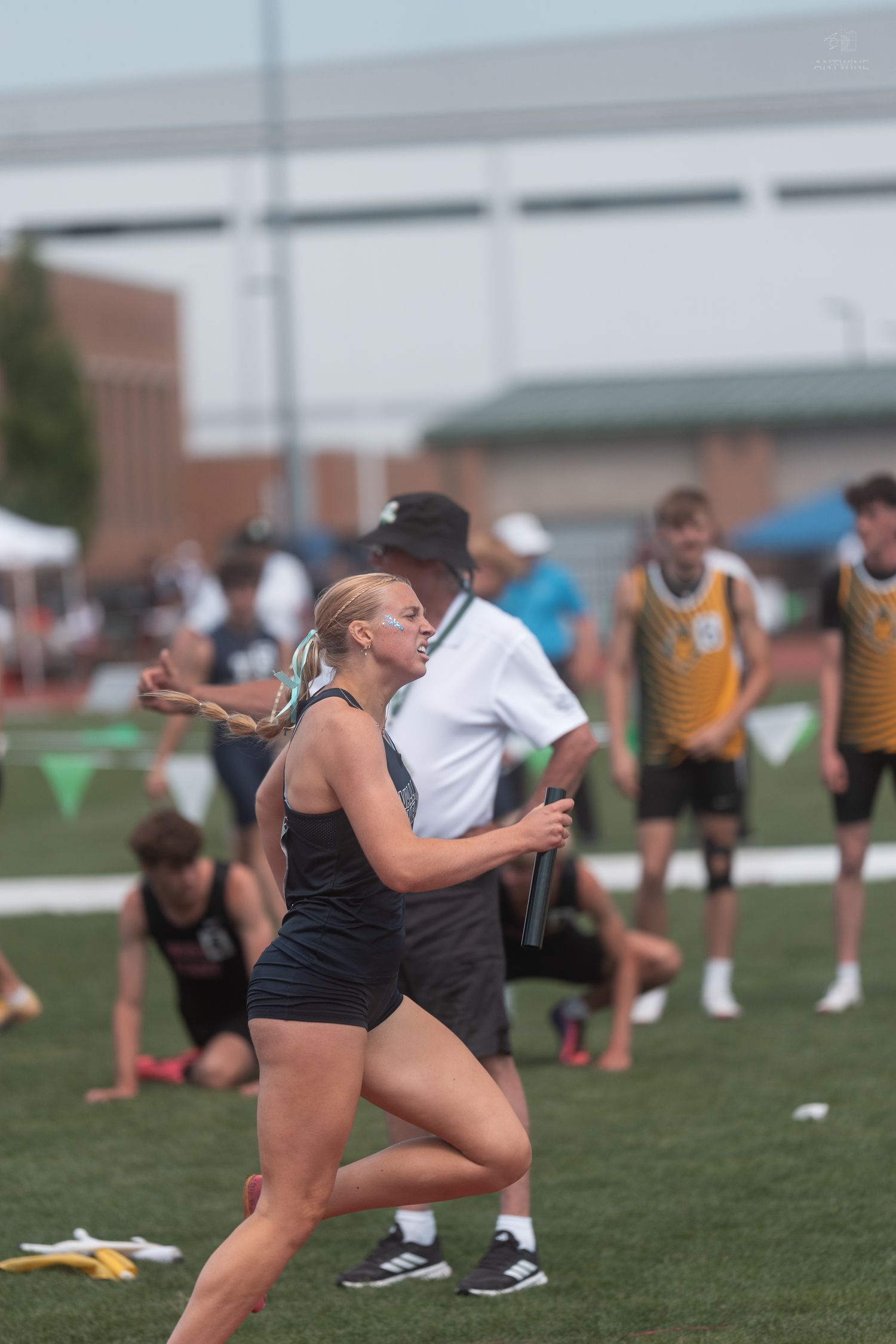 Blonde female sprinter in black athletic uniform accelerates during relay race at outdoor track event.