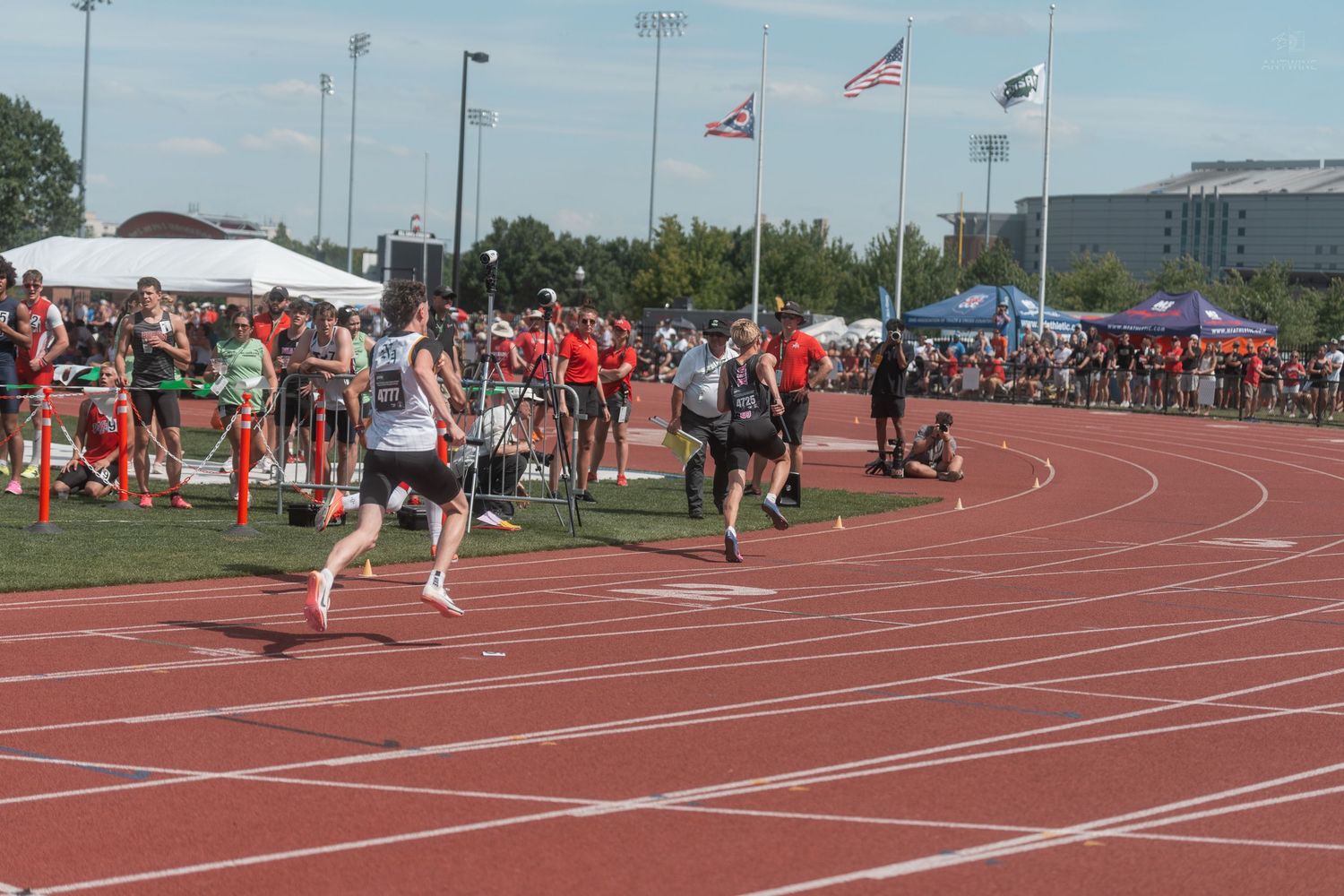 A lone runner races on a red track at a high school track and field meet with flags flying overhead.