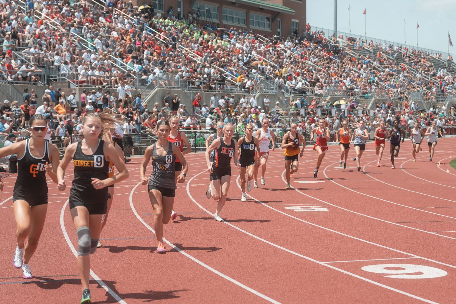 Female runners compete in a track race at a large high school track and field meet with thousands of spectators.