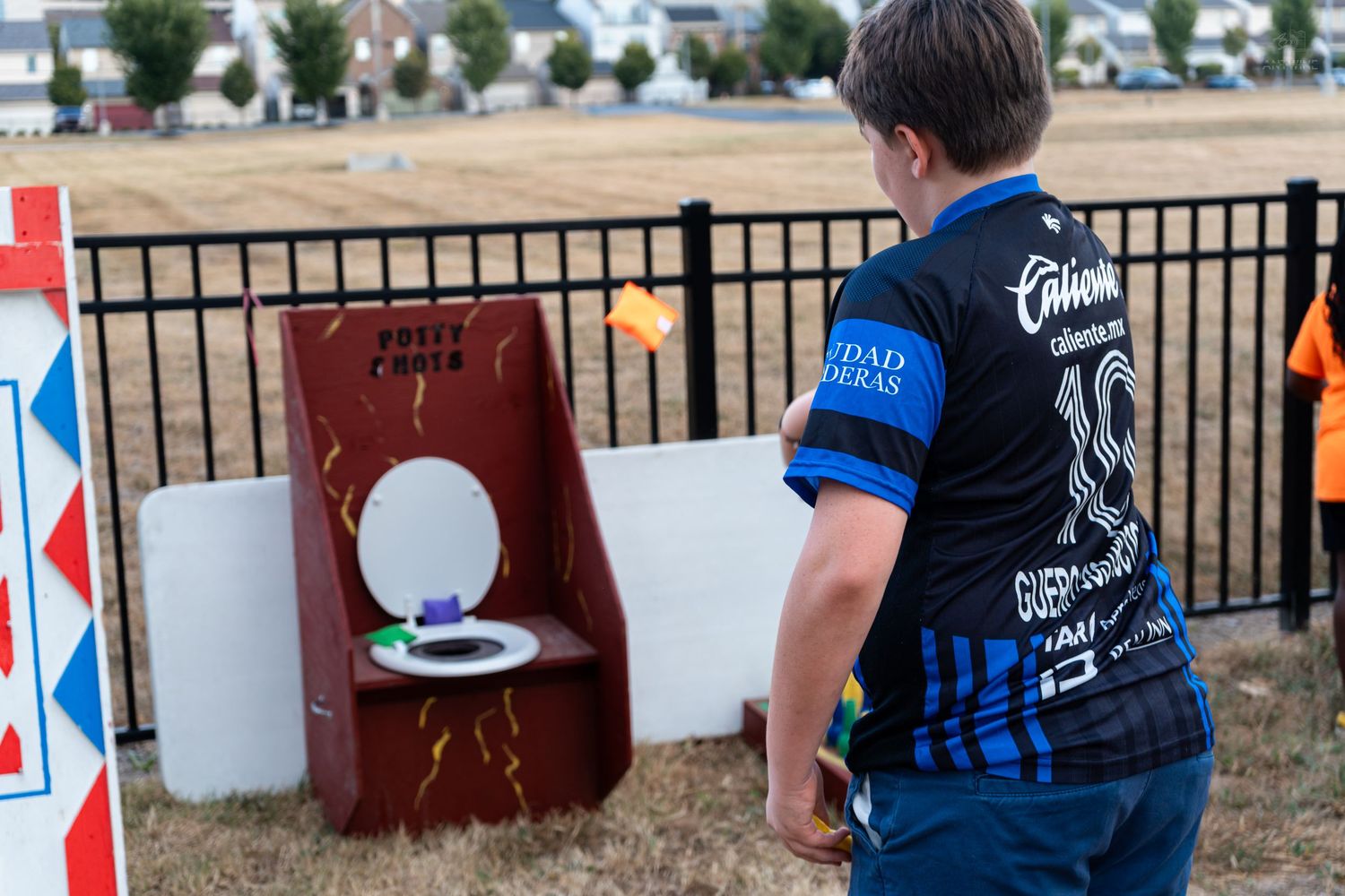 Child plays potty-themed cornhole toss game at outdoor carnival fair event wearing blue soccer jersey.