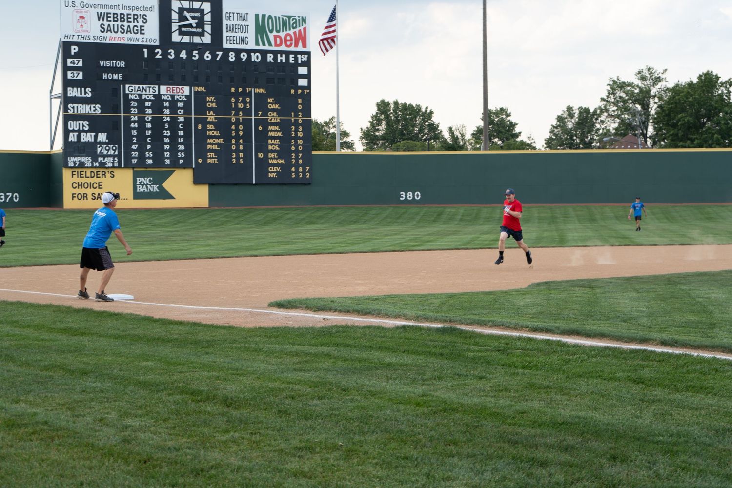 Players practice on a green baseball field with a large scoreboard and Mountain Dew advertisement in the background.