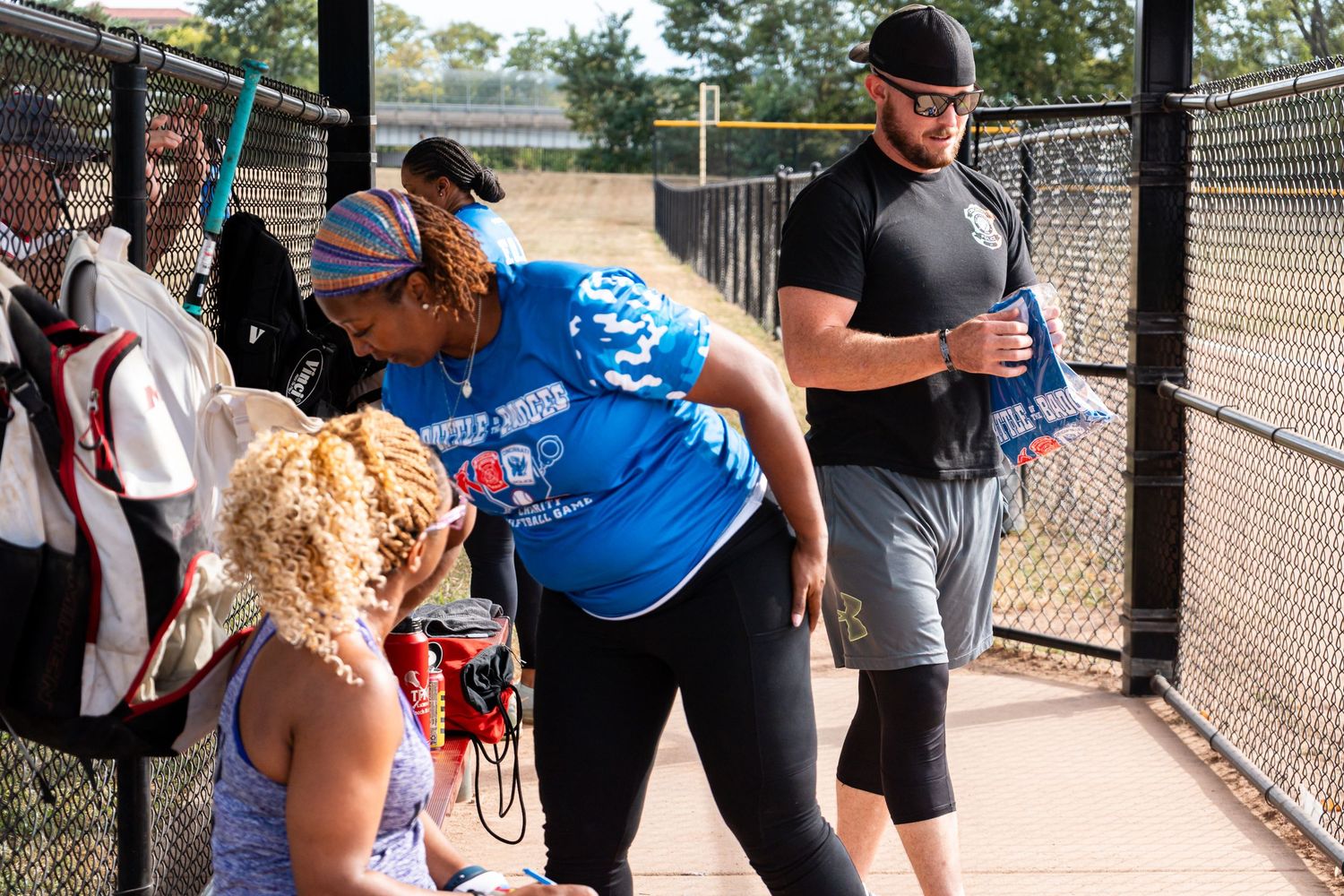 People gathering at a baseball field fence, wearing casual sportswear on a sunny day outdoors.