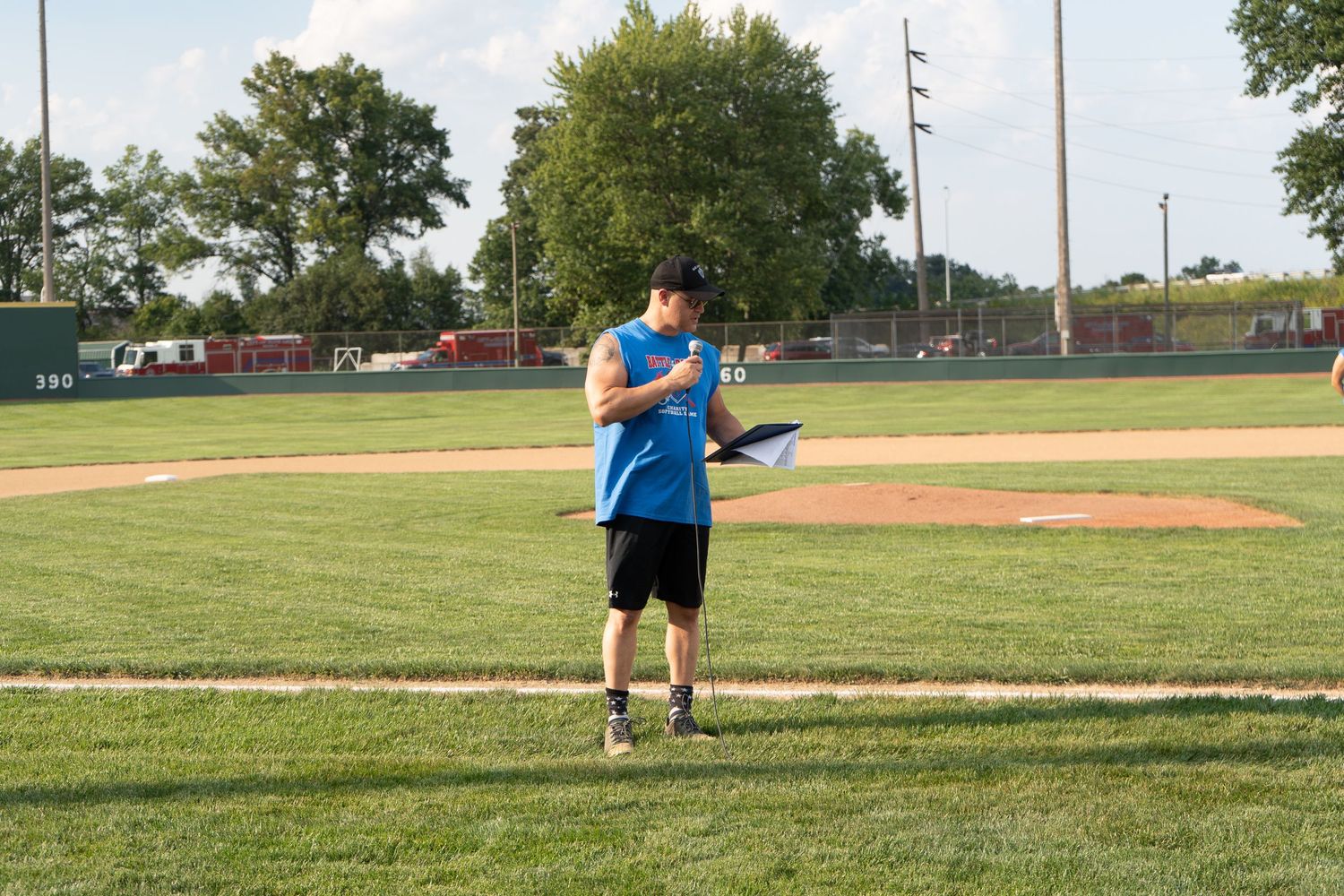 A coach in a blue jersey reviews notes on a clipboard while standing on a green baseball field on a sunny day.