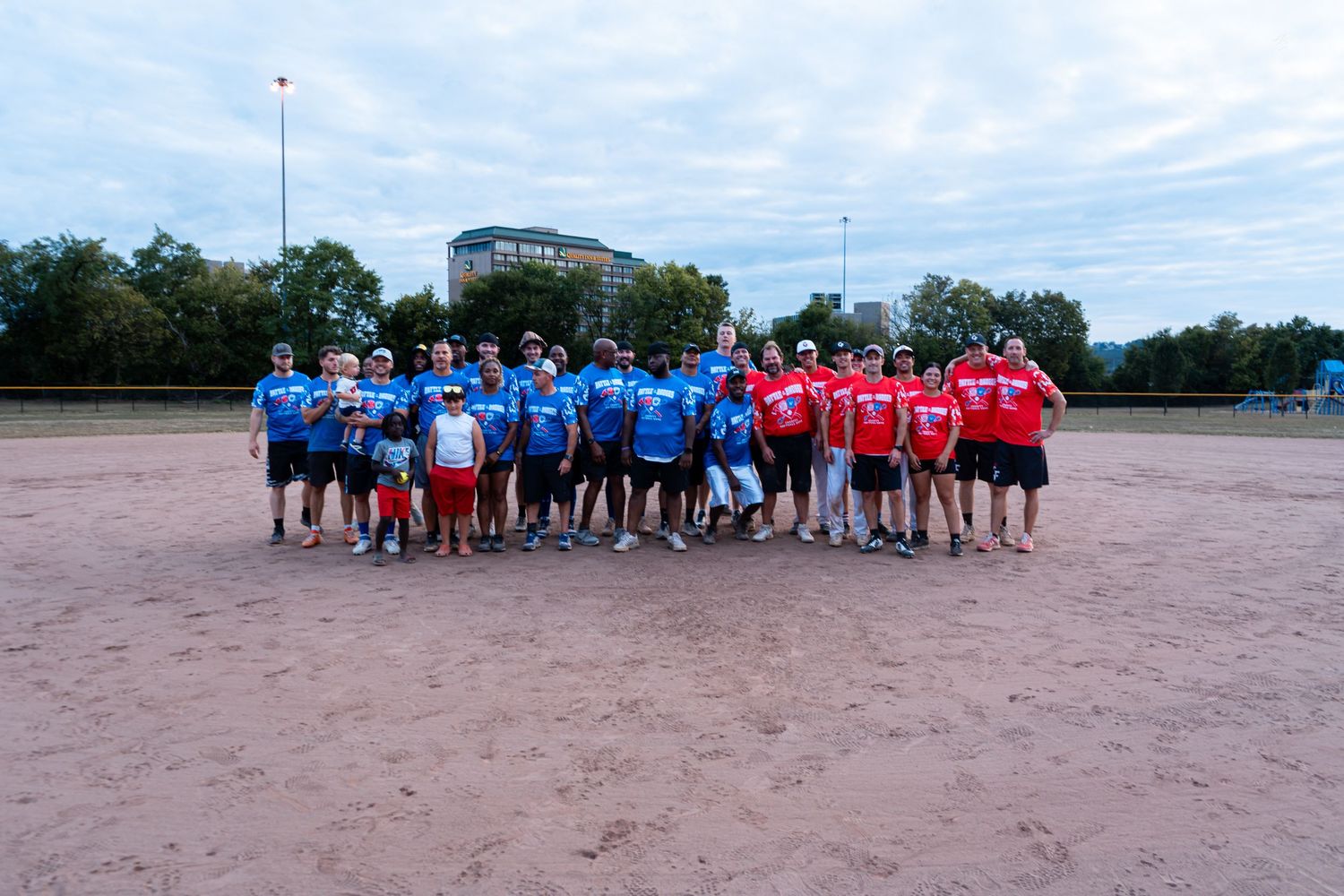 Two softball teams in red and blue jerseys pose together on a dirt field under cloudy skies.