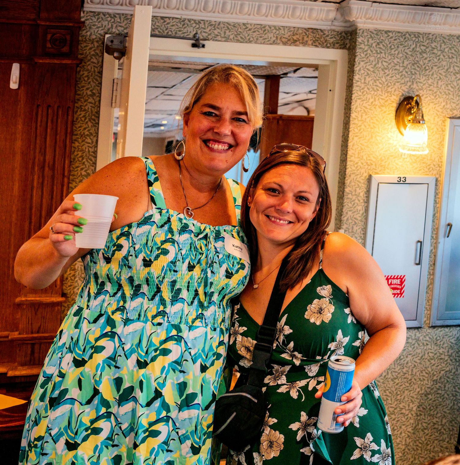 Two smiling women pose together at an indoor event, one in a floral dress holding a cup, the other in a green dress.