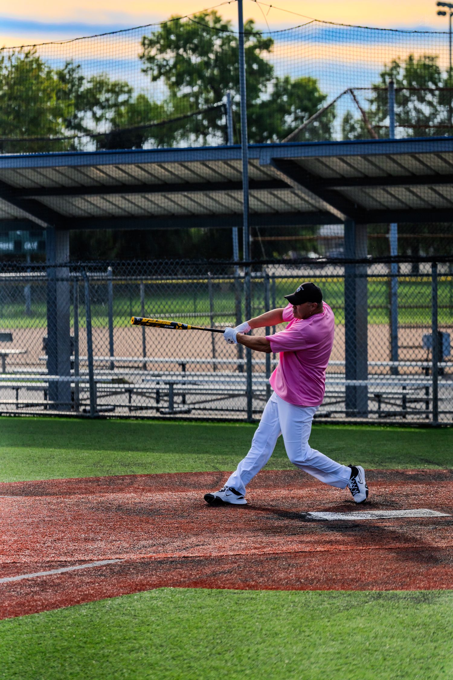 A batter in a pink shirt and white pants swings a baseball bat at home plate during a sunset game.