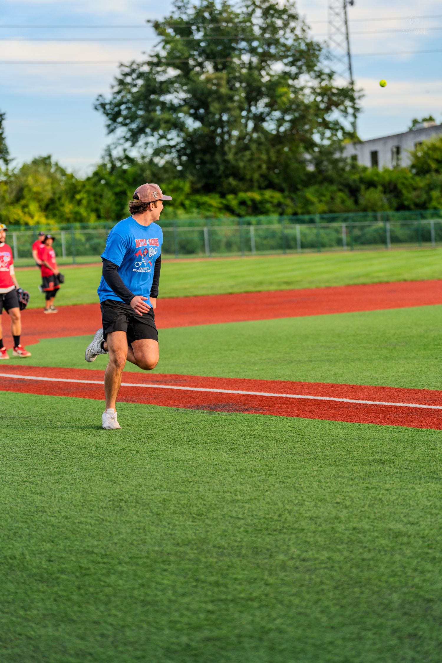 A man in a blue shirt and cap runs across a green turf baseball field during an outdoor softball game.