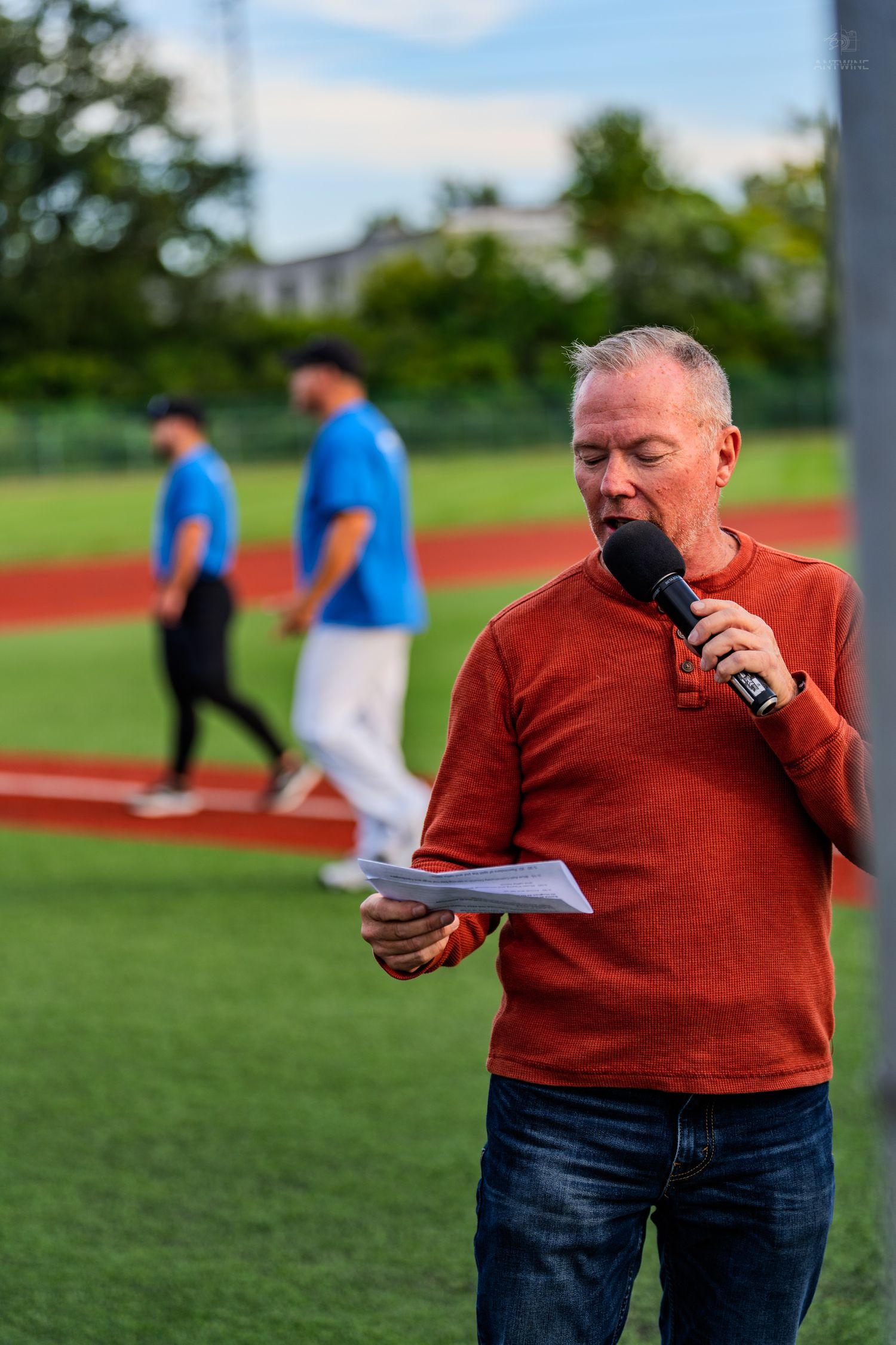 Sports announcer in red sweater reading notes into microphone at baseball field.