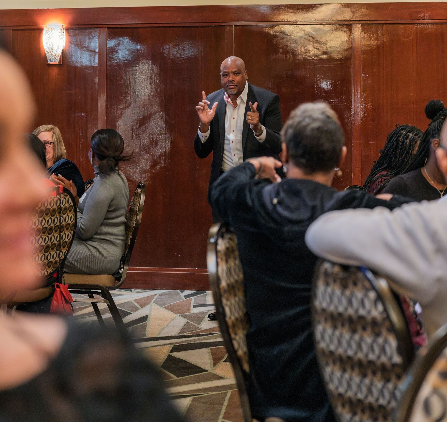 Man in suit speaking animatedly to seated audience in wood-paneled event room.