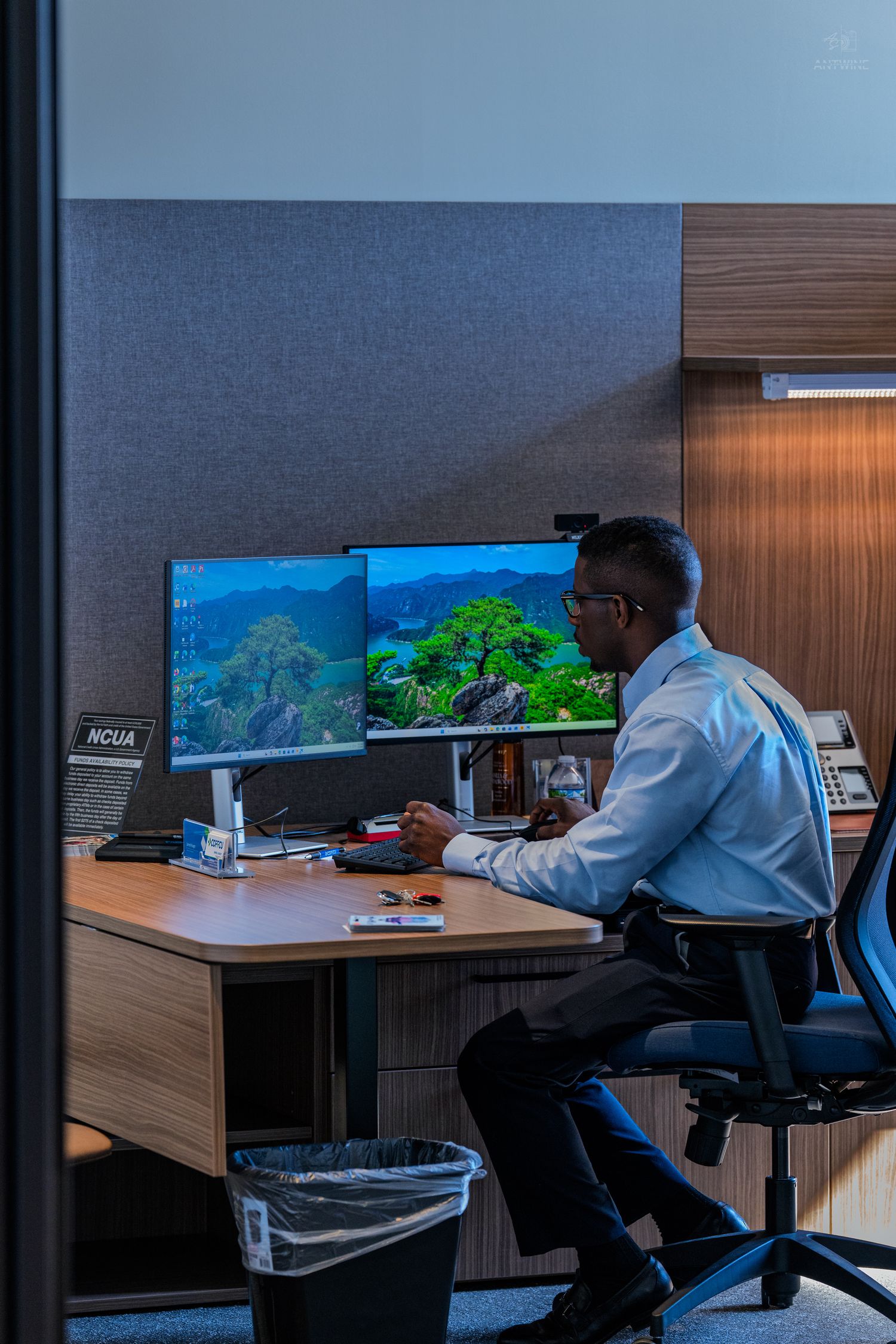 A Black professional man in a white shirt works at a dual-monitor desk setup in a modern office cubicle.