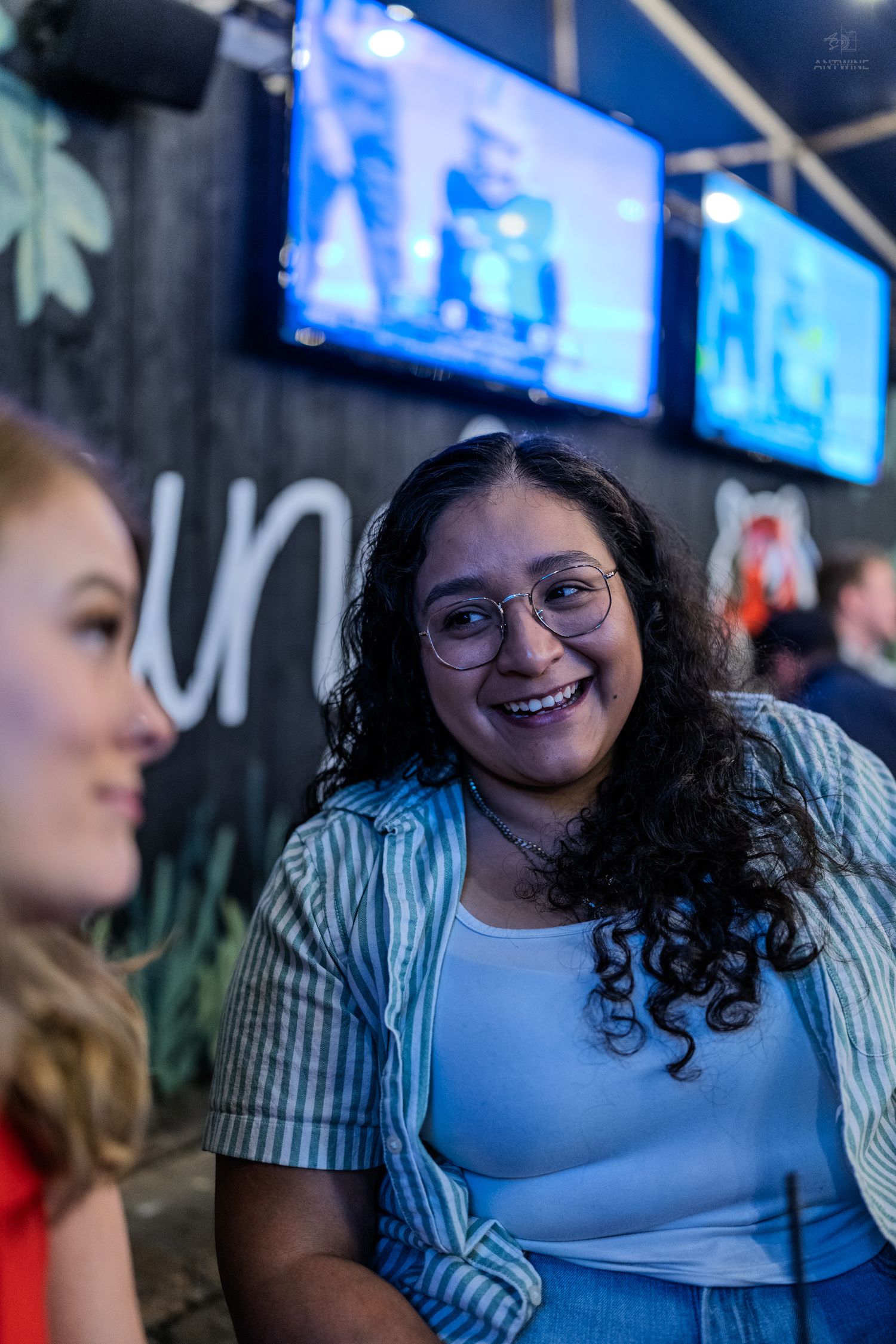 Two young women smiling and chatting at a social event with TV screens in the background.