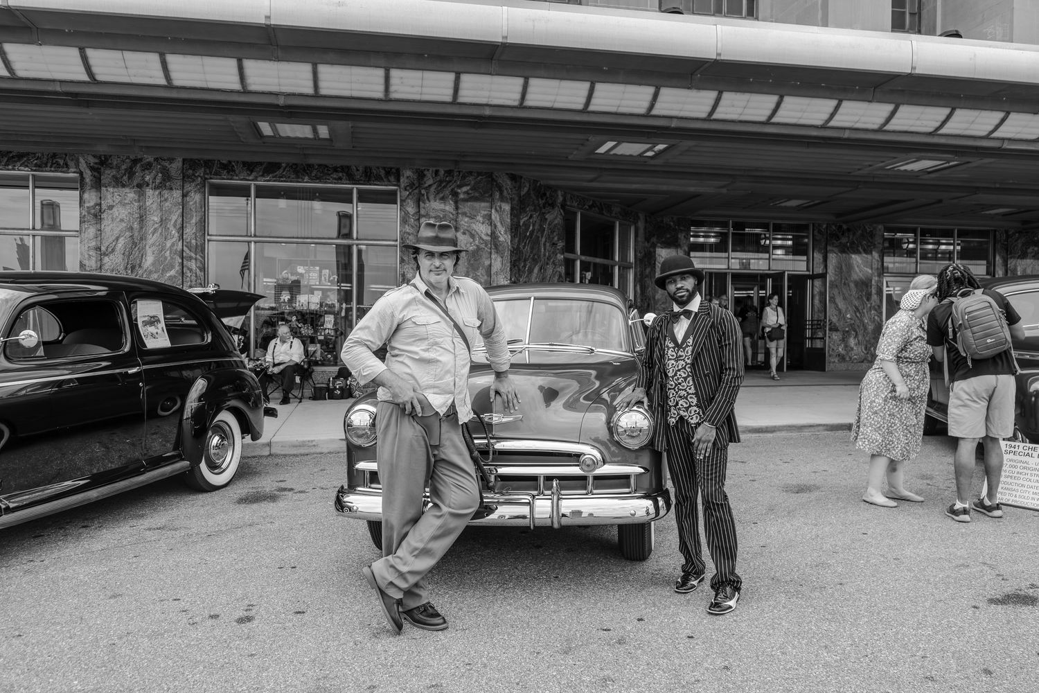 Two men pose beside a vintage 1950s car outside a building in this classic black and white photograph.