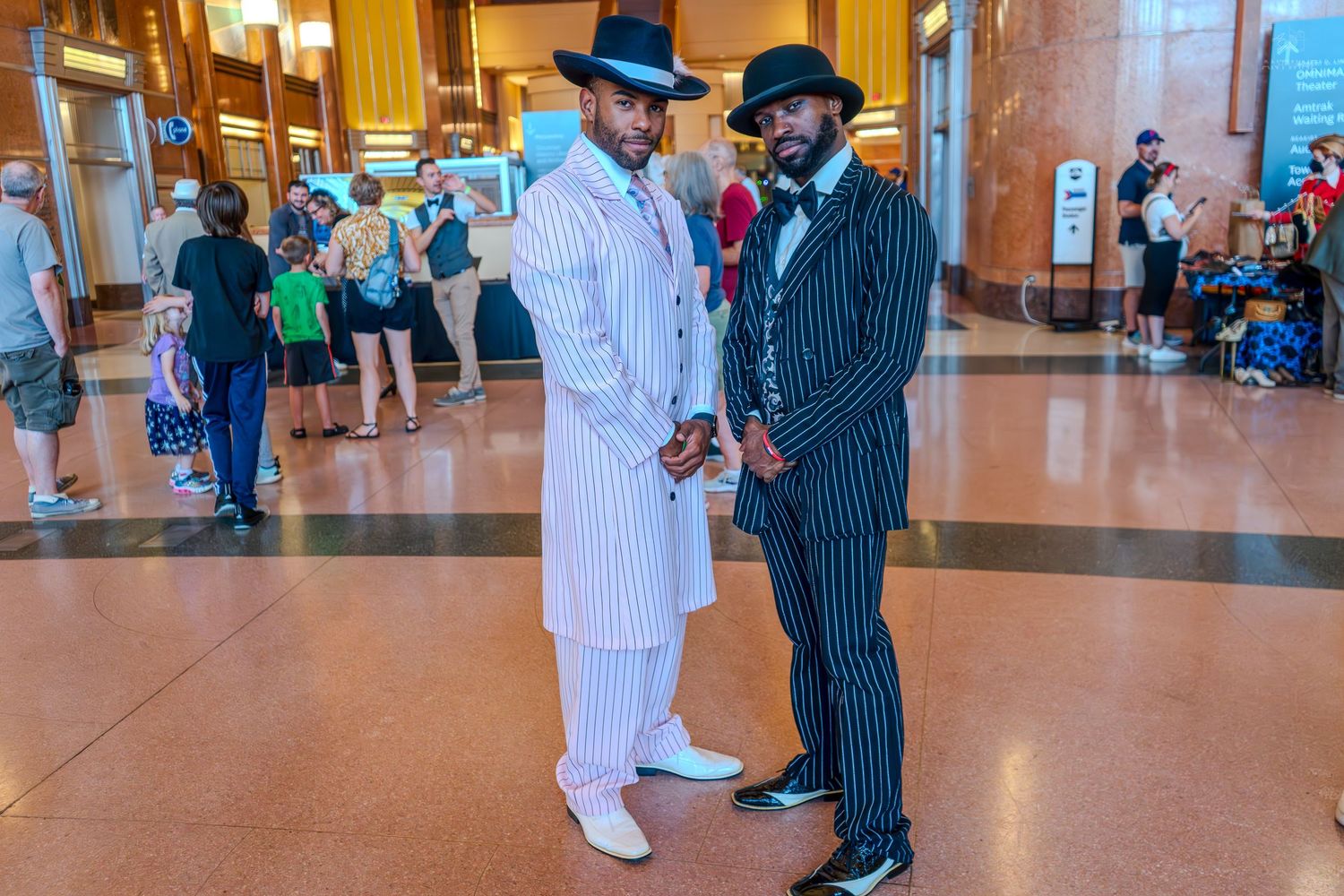 Two men in pinstripe zoot suits and fedora hats pose in a grand building interior.