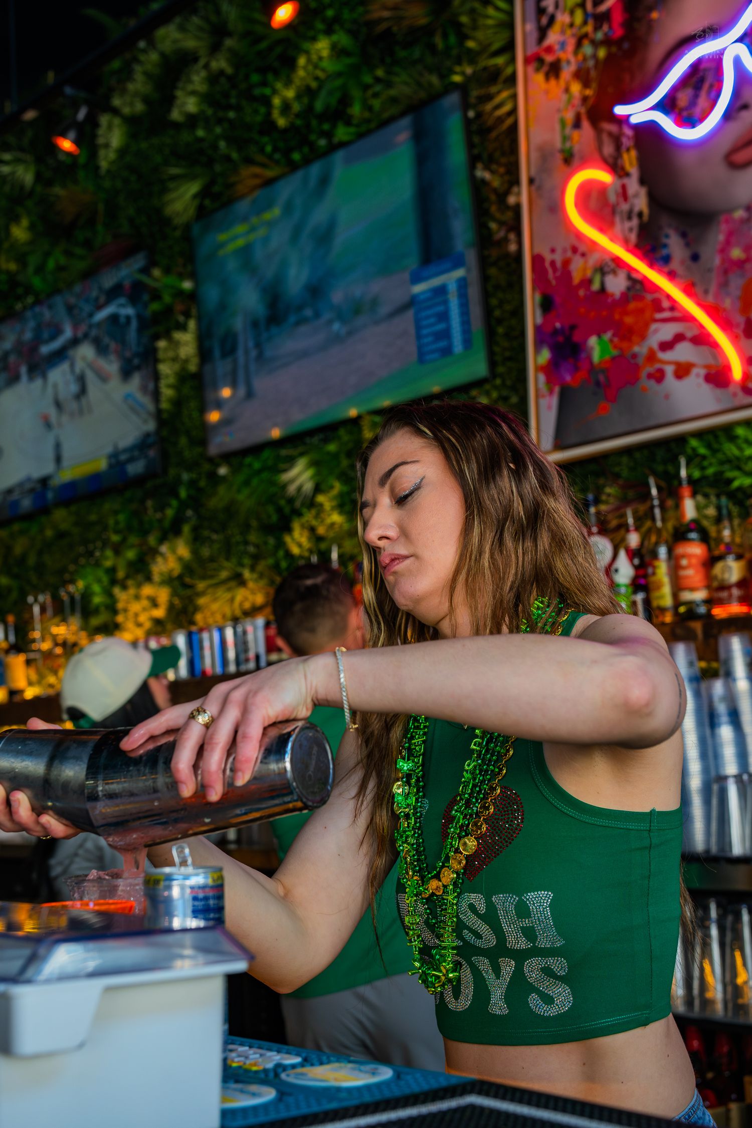 Bartender in green Irish Boys top pouring cocktail shaker at a vibrant bar with neon lights and TVs.