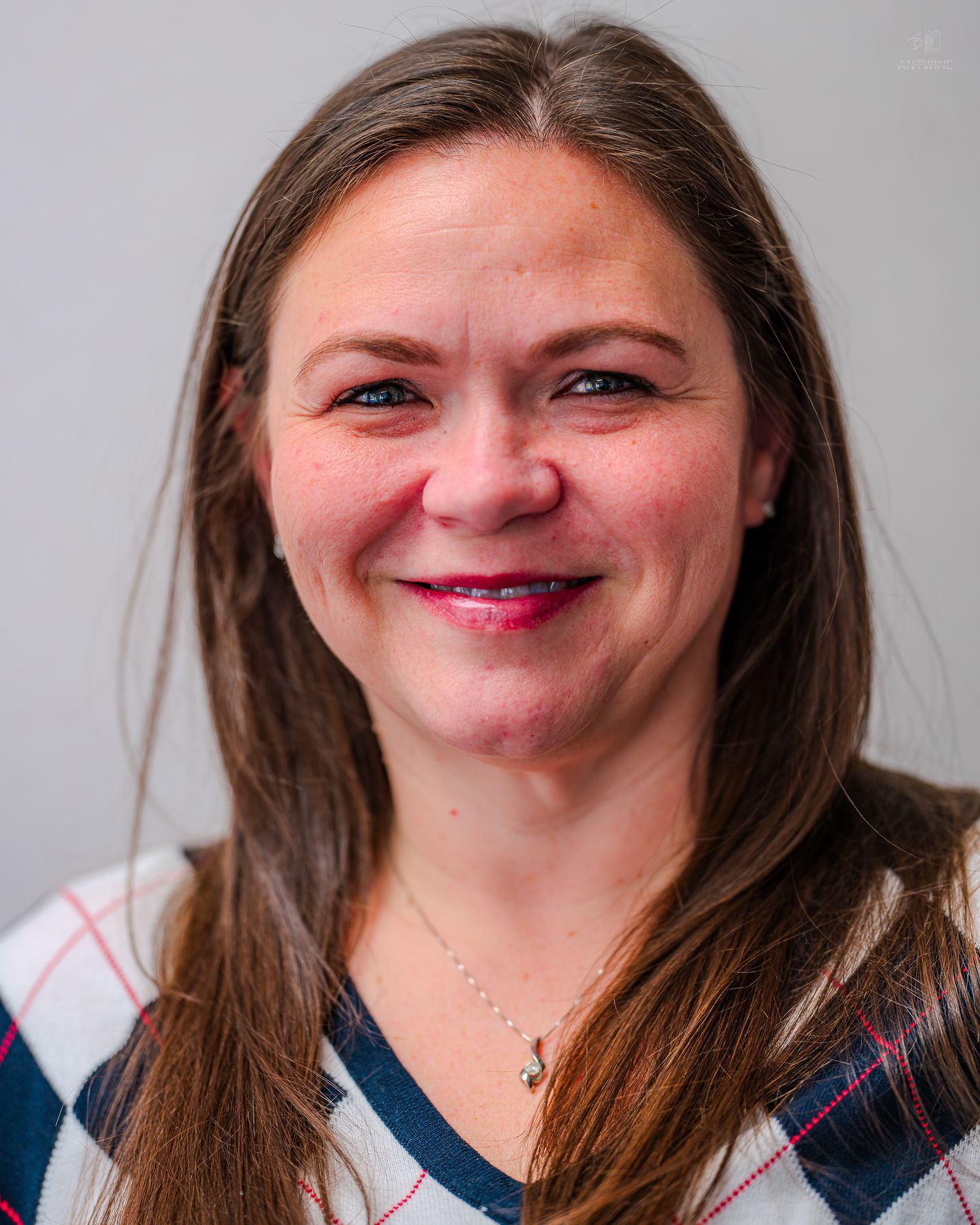Professional headshot of a smiling woman with long brown hair wearing a plaid top and delicate necklace.