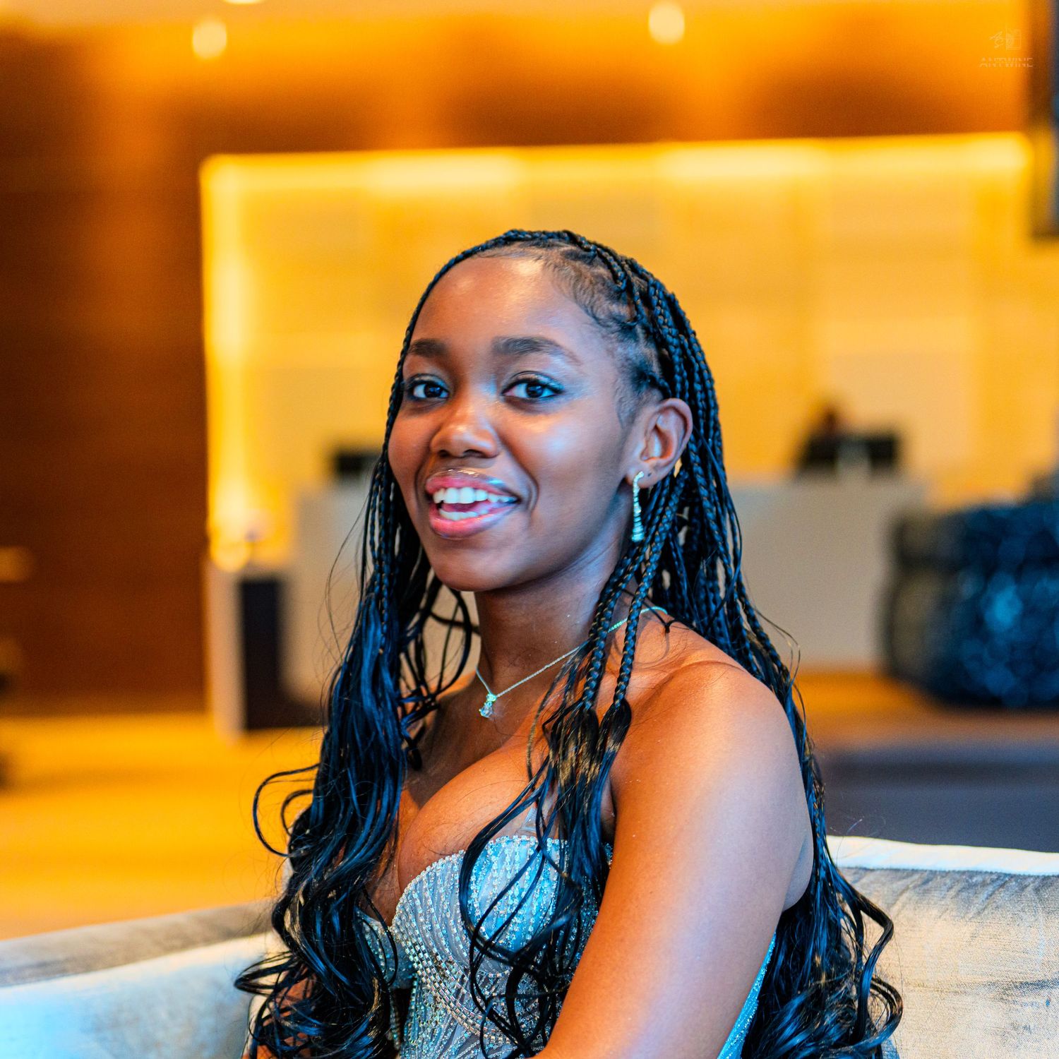 Young woman with long braids smiling in elegant attire at a modern hotel lobby.