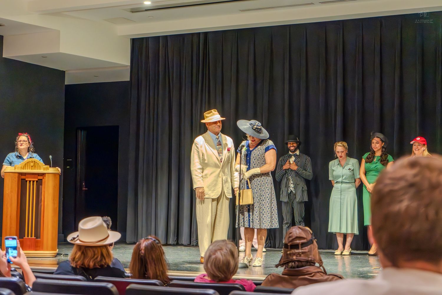 Actors perform a vintage-themed stage play in a theater, wearing 1940s costumes including suits and dresses.