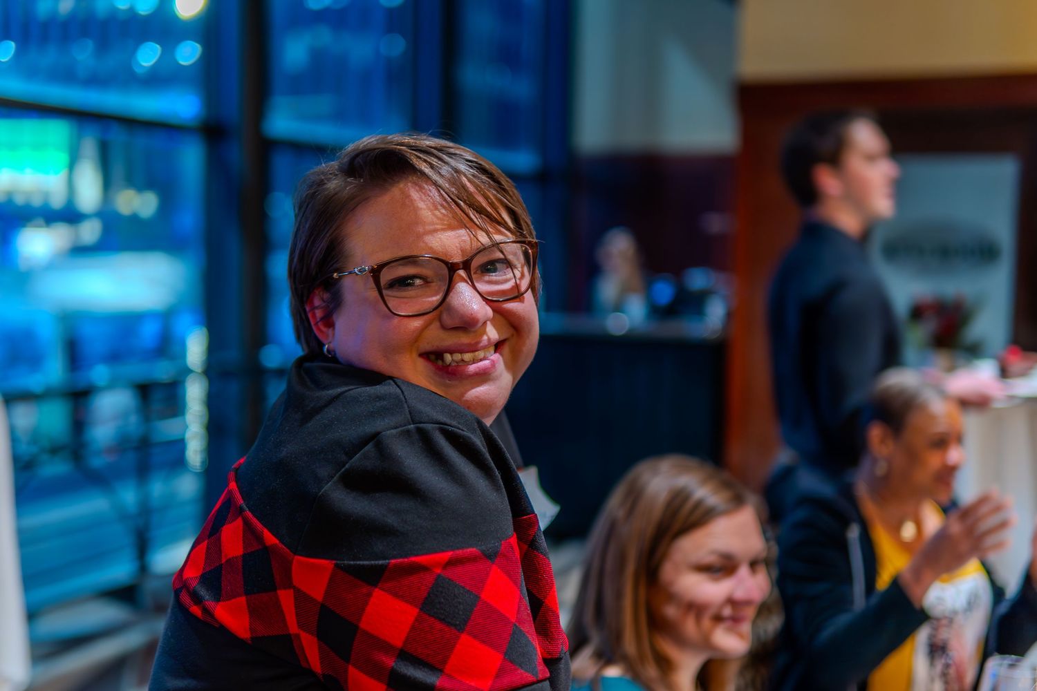 Smiling person with glasses and red plaid jacket at an indoor event with people in background.