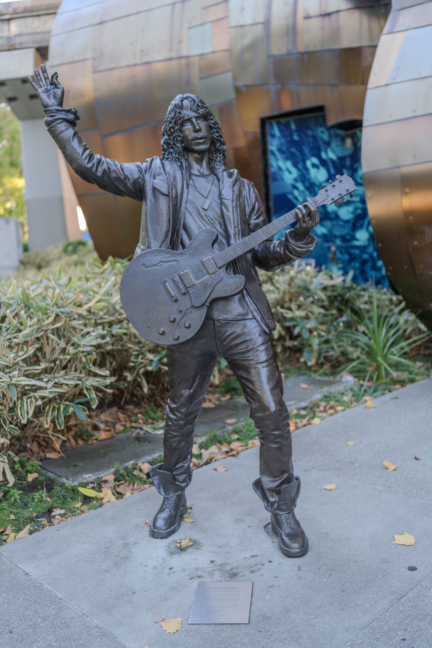 Bronze statue of a rock guitarist with one arm raised outside a modern building.
