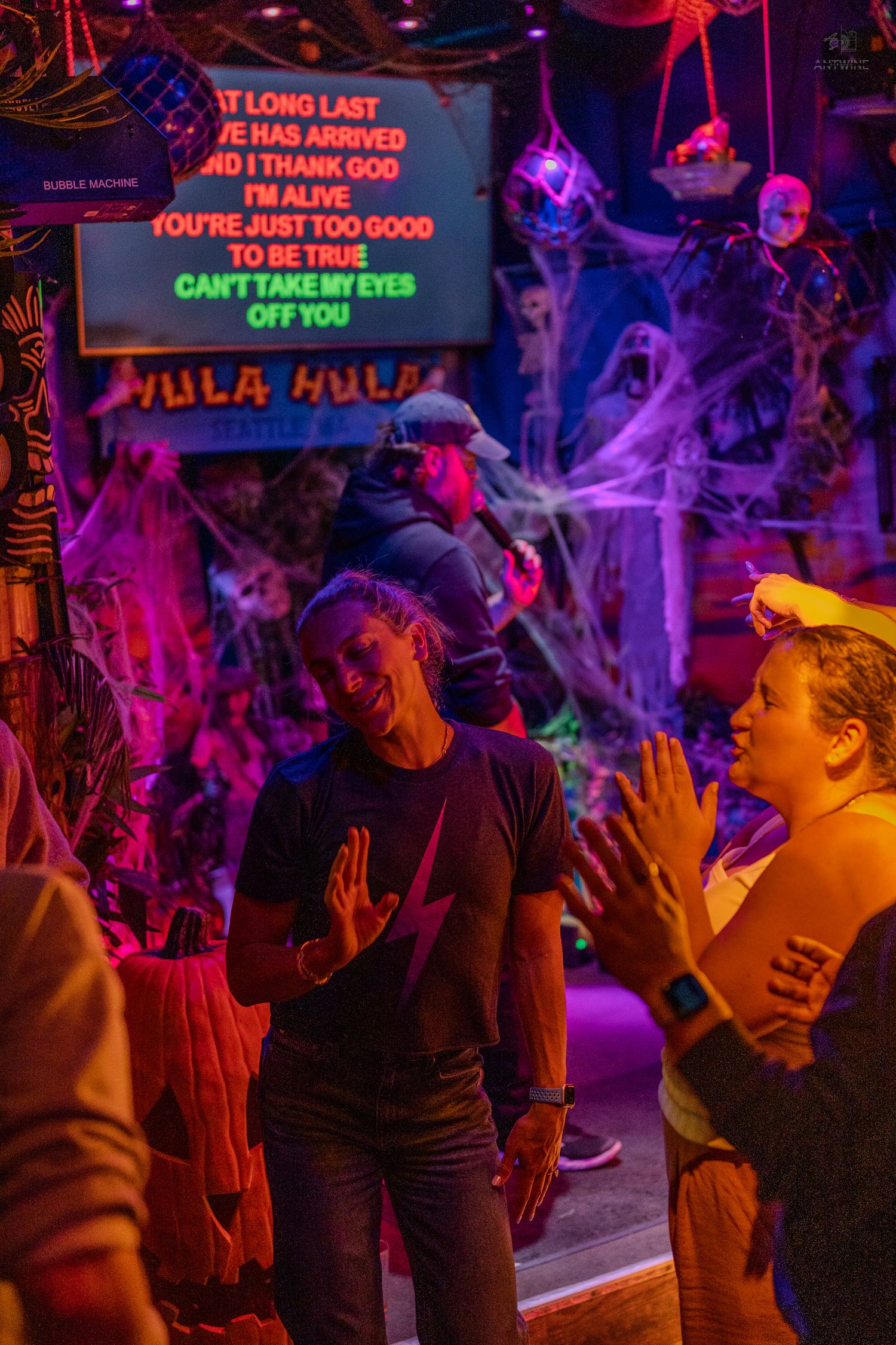People enjoying Halloween karaoke at neon-lit Hula Hula bar with spooky cobweb decorations.