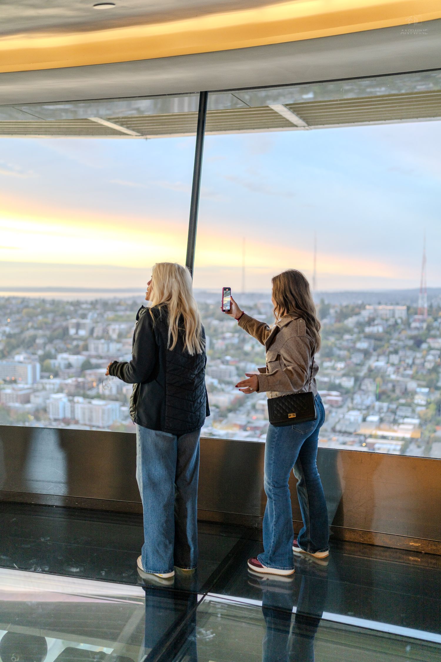 Two women stand at floor-to-ceiling windows of a high-rise observation deck, one taking a photo of the city skyline at sunset.