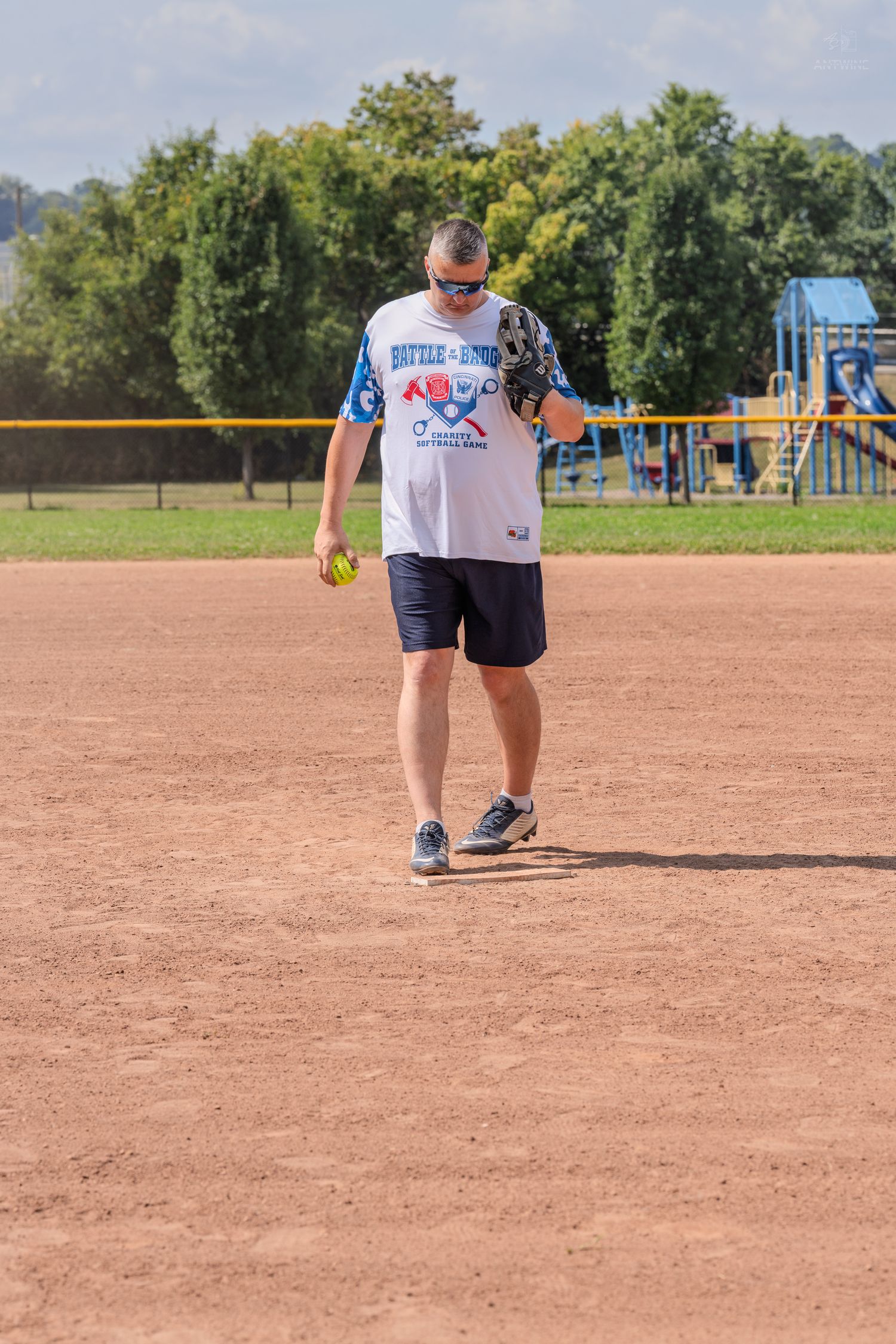 Male softball player walking on a dirt baseball field holding a yellow ball and wearing a glove.
