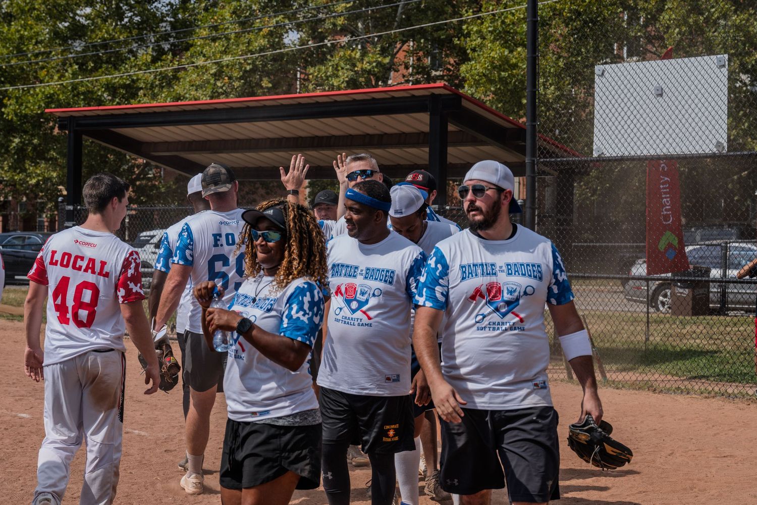 Softball players in Battle of the Badges charity game uniforms celebrate on the field on a sunny day.