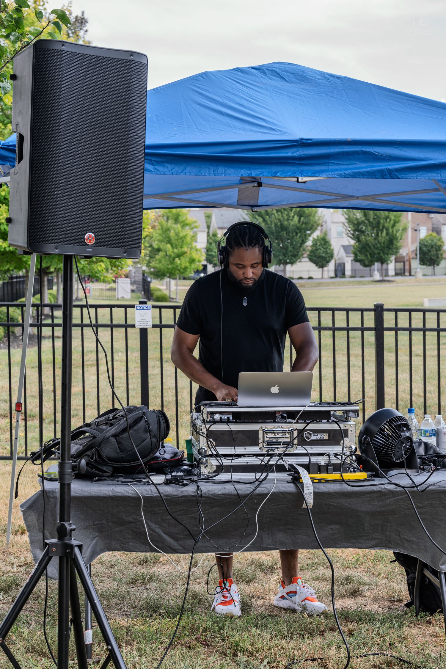 Outdoor DJ in black outfit operates laptop and speaker setup under blue tent.