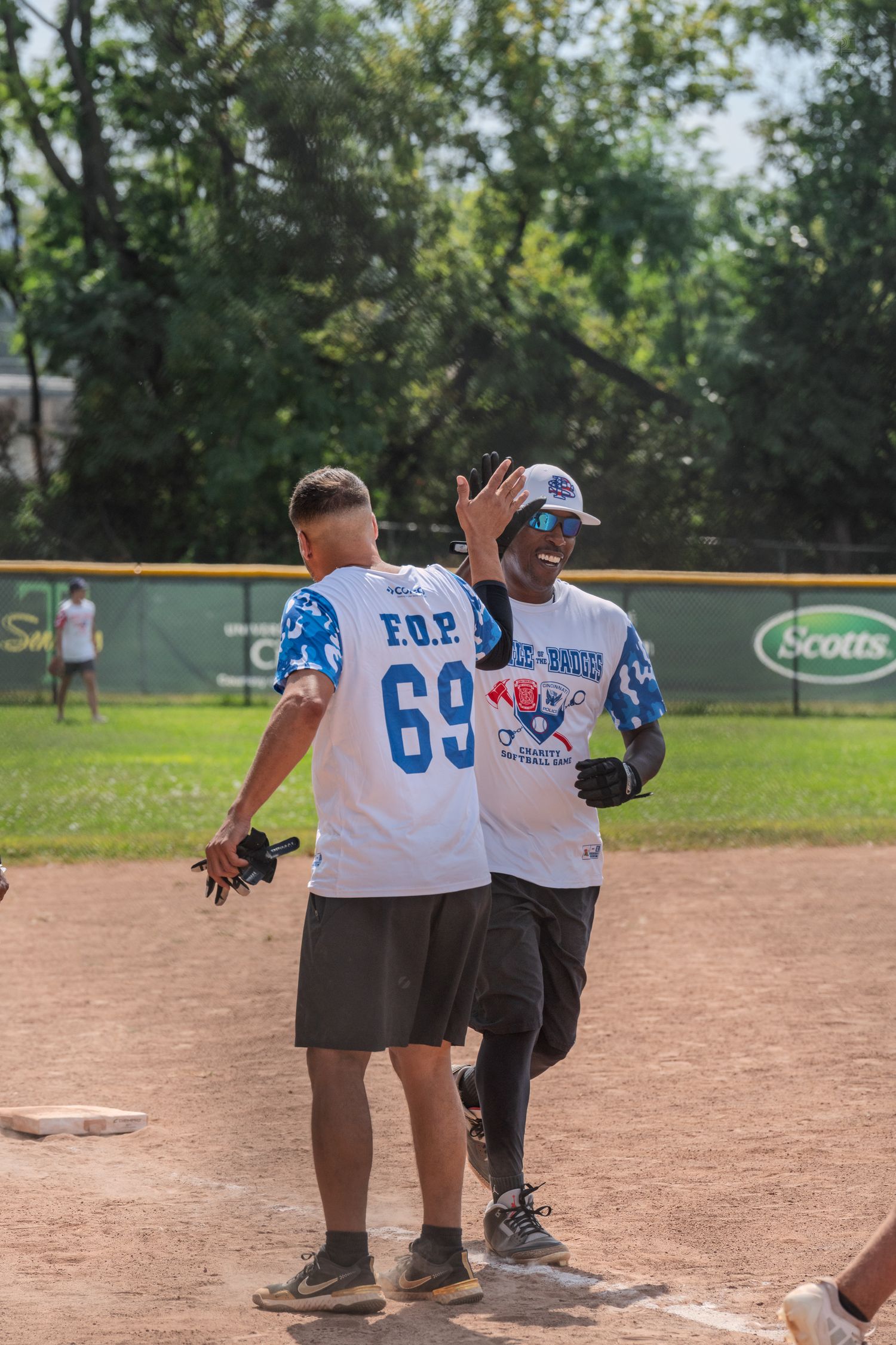 Two softball players in FOP jerseys celebrate with a high five on the baseball diamond during a charity game.